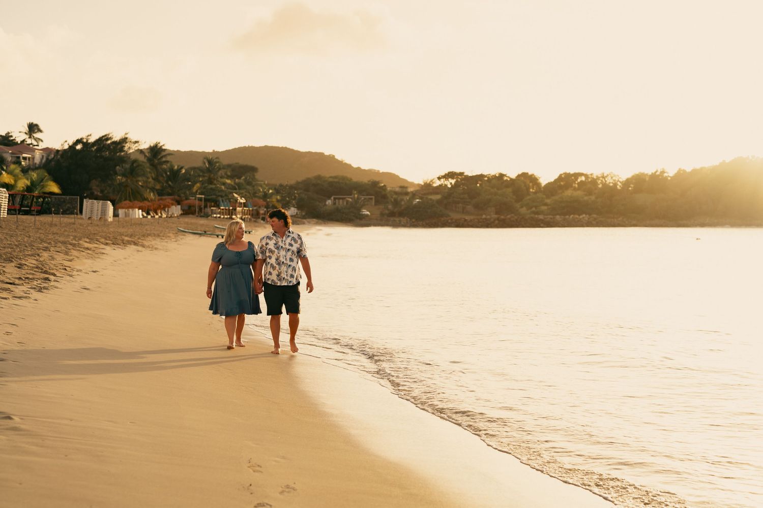 Couple walking hand in hand along shoreline during warm golden sunset on beach.