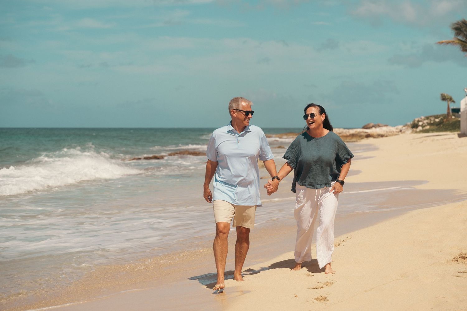 A mature couple in casual attire walks hand-in-hand along a sunny tropical beach with ocean waves.