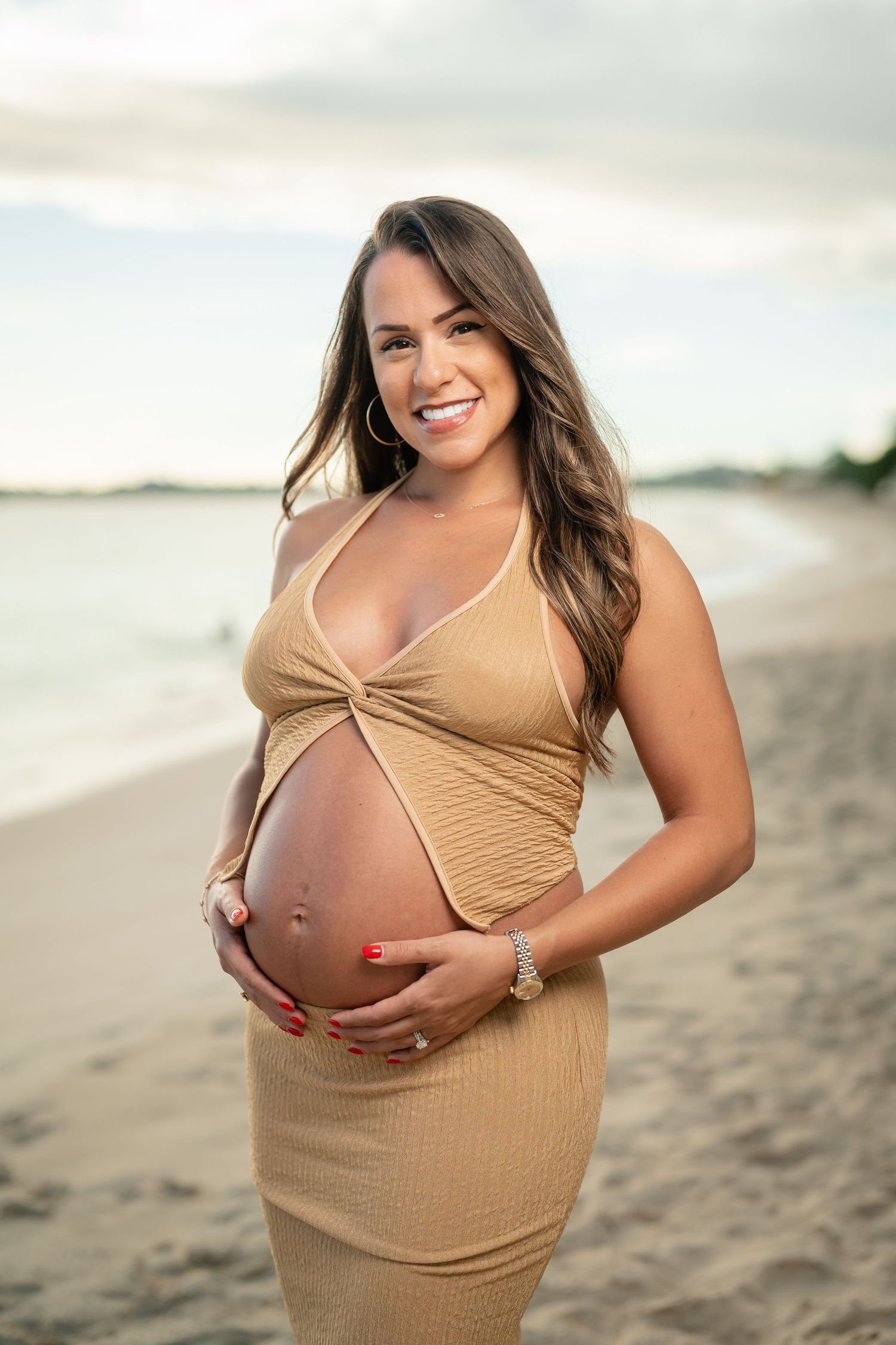 Smiling pregnant woman in gold dress cradling baby bump on beach at sunset.