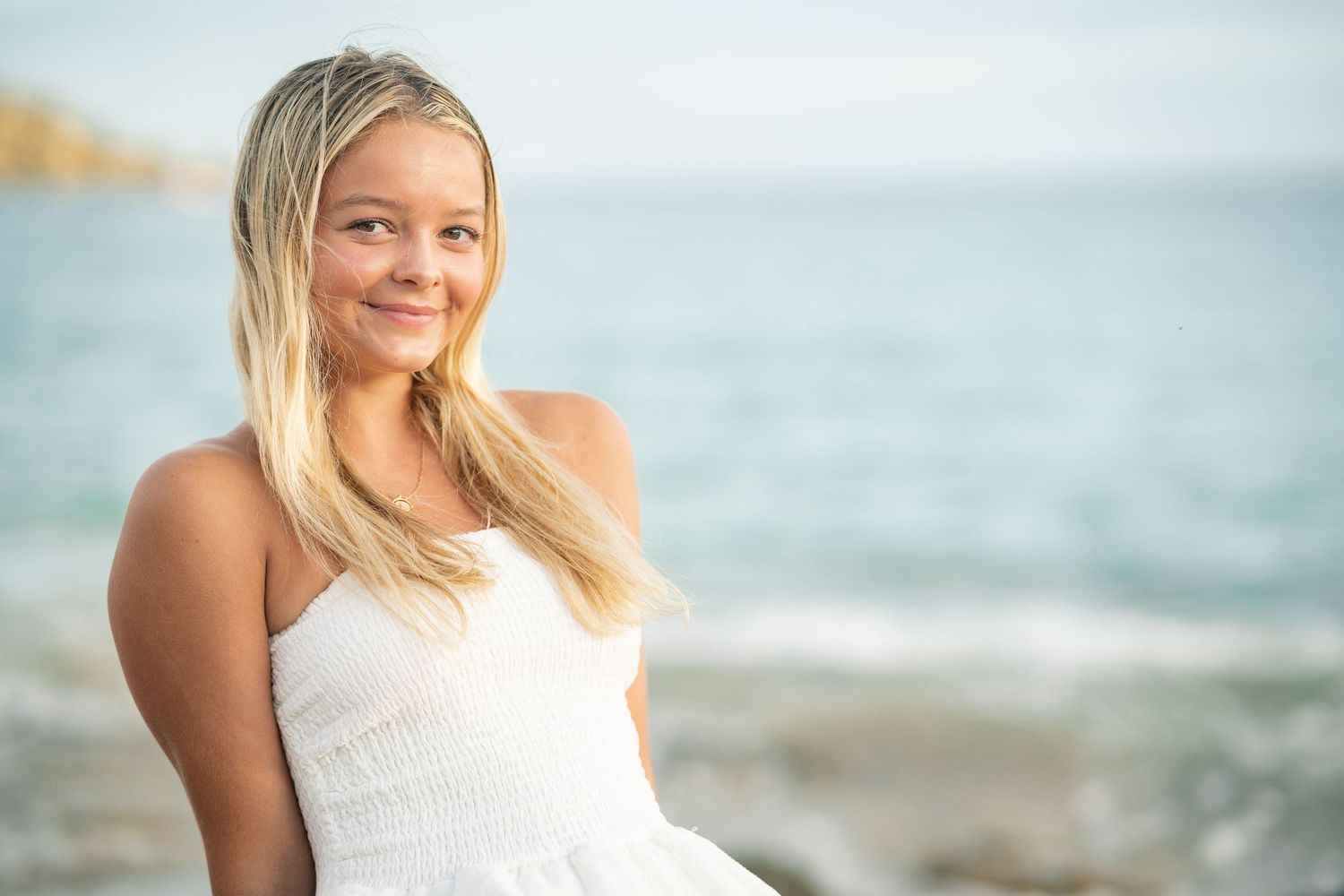 Smiling blonde teenage girl in white sundress sitting on rocks with blue ocean in background.