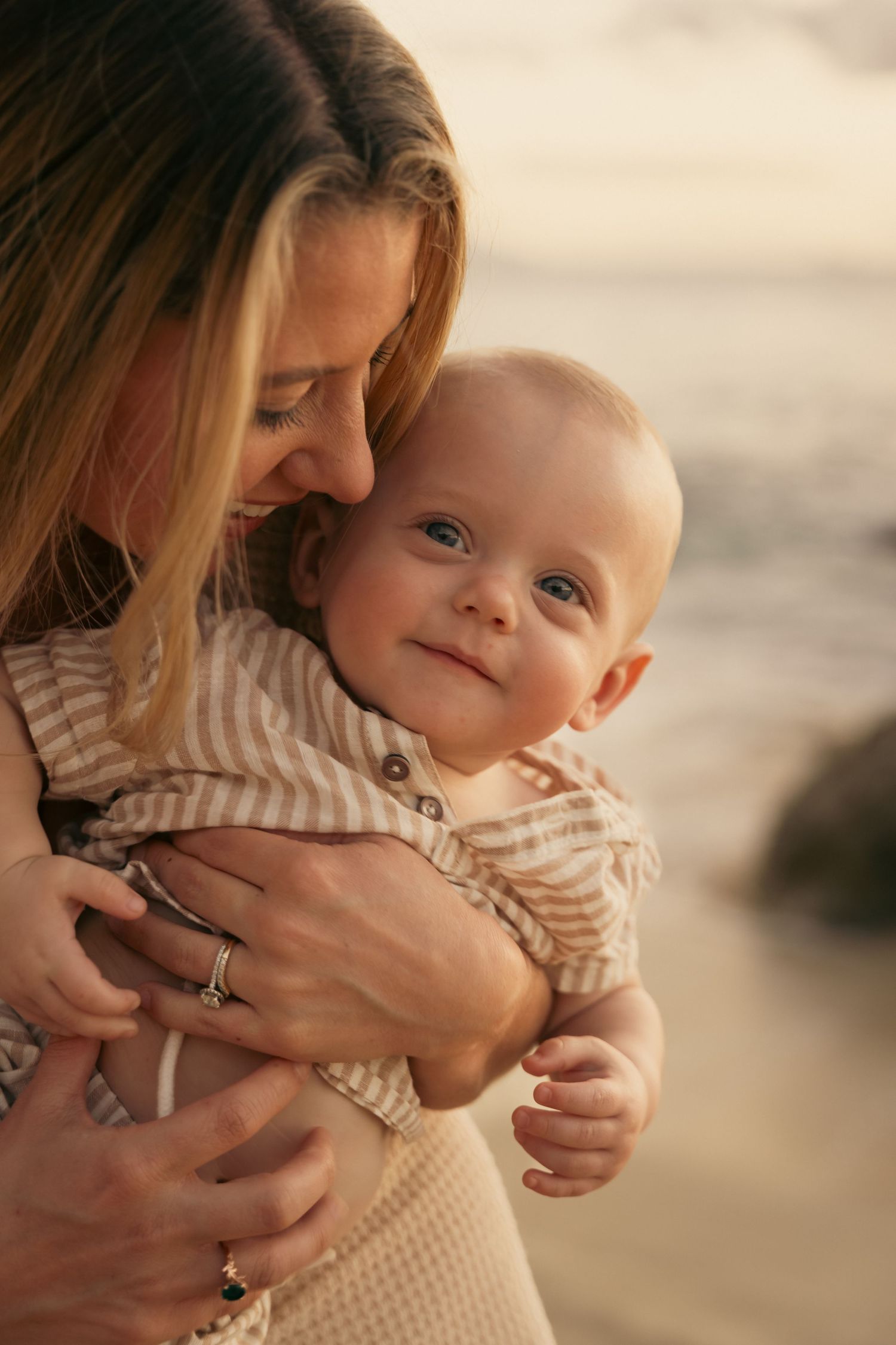 Mother tenderly holding smiling baby near the ocean at golden hour.