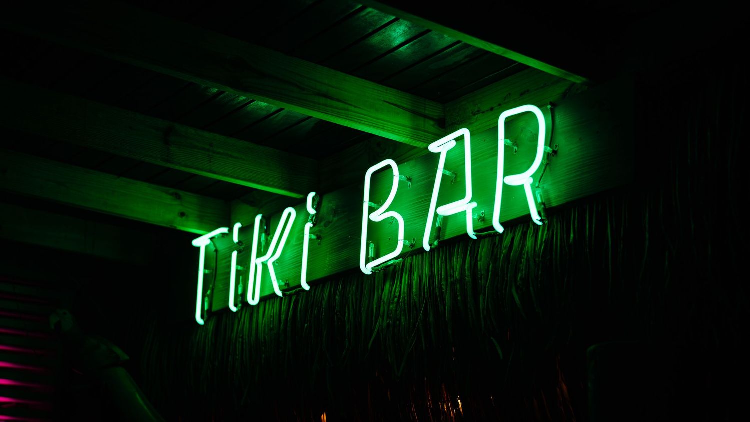 Green neon Tiki Bar sign glowing in the dark against a wooden ceiling backdrop.