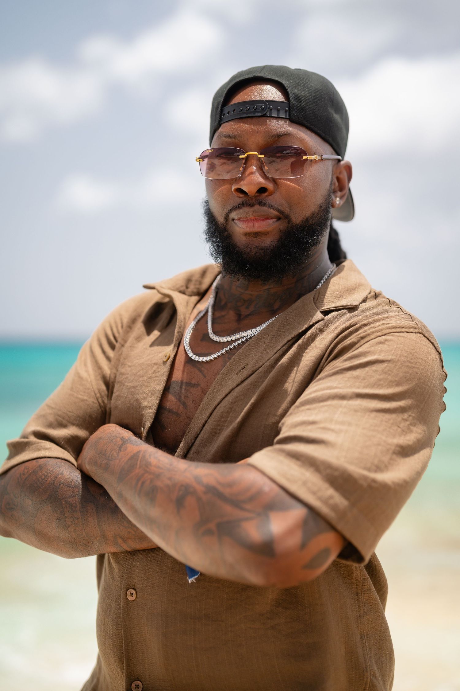 Tattooed bearded man in brown shirt, cap, and sunglasses stands with arms crossed on a tropical beach.