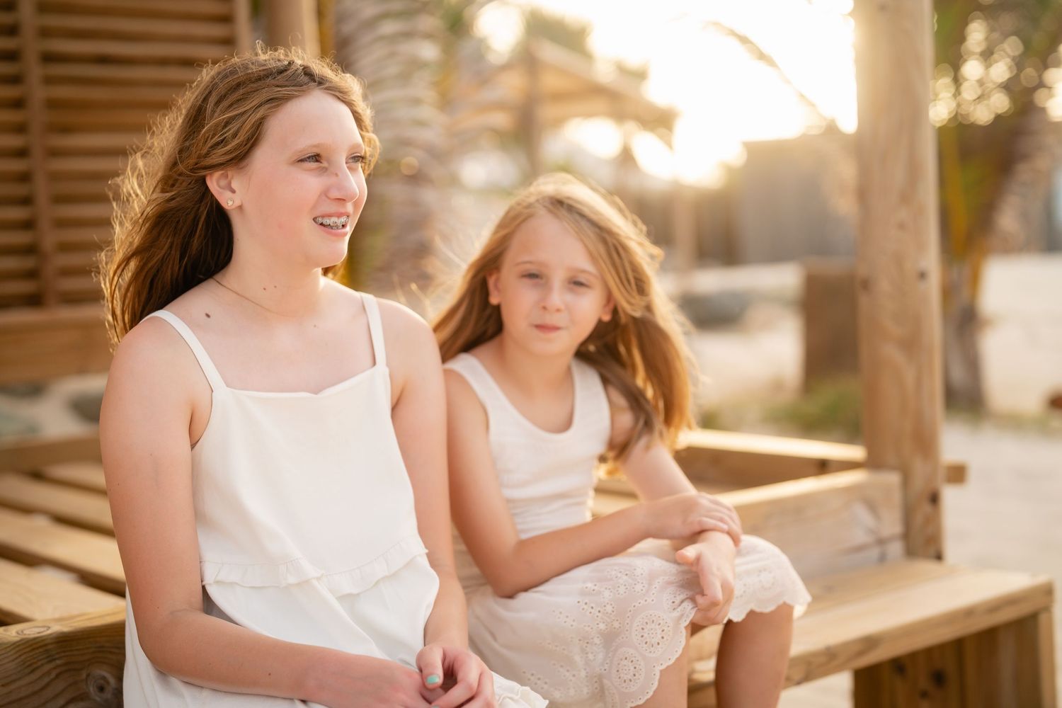 Two sisters in white dresses sitting together on wooden steps in golden sunlight.