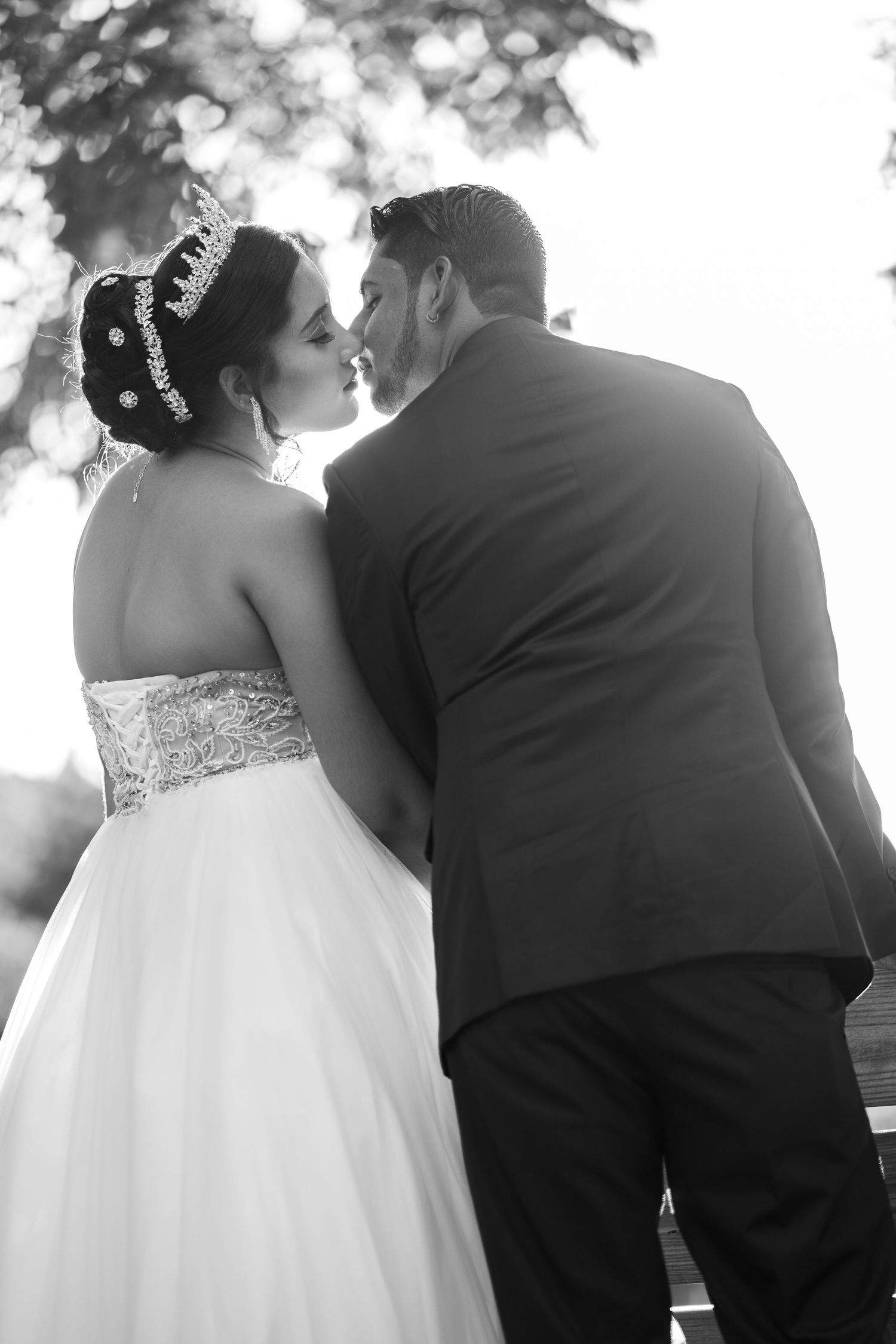Bride in white ball gown with tiara and groom in black suit share a kiss outdoors.