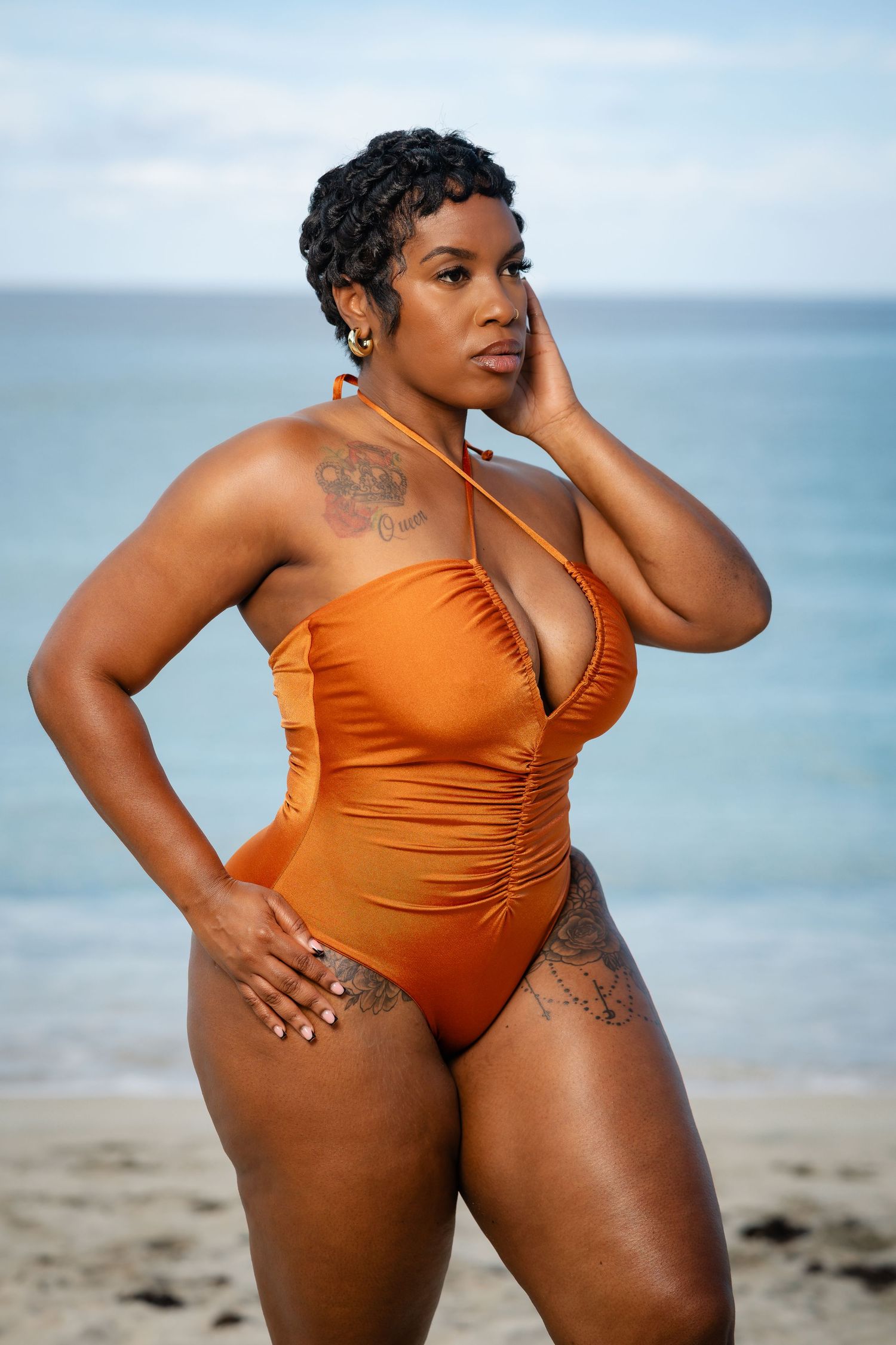 Confident plus-size woman in orange swimsuit posing on a beach with ocean backdrop.