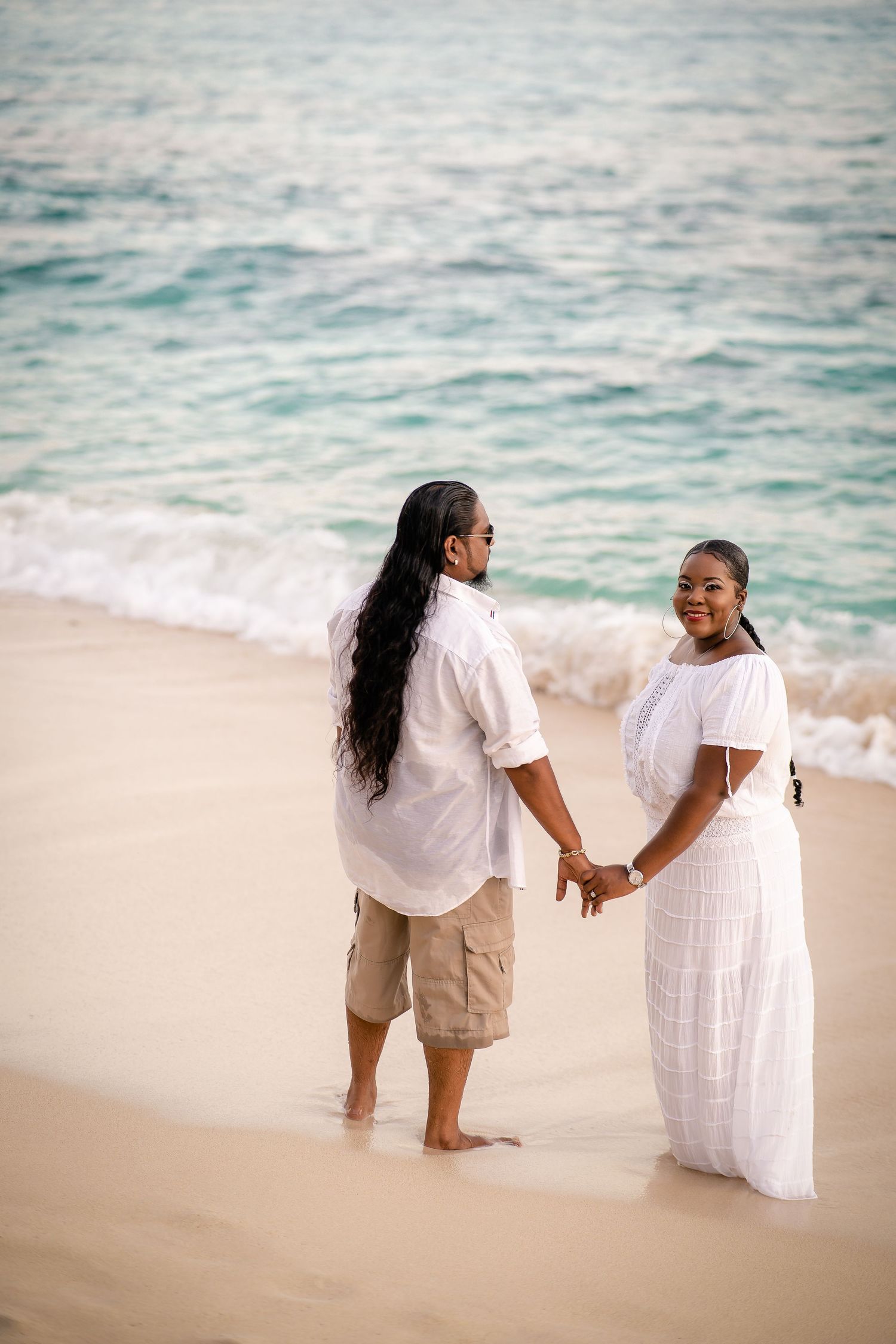 Couple in white outfits holding hands on sandy beach with turquoise ocean waves.