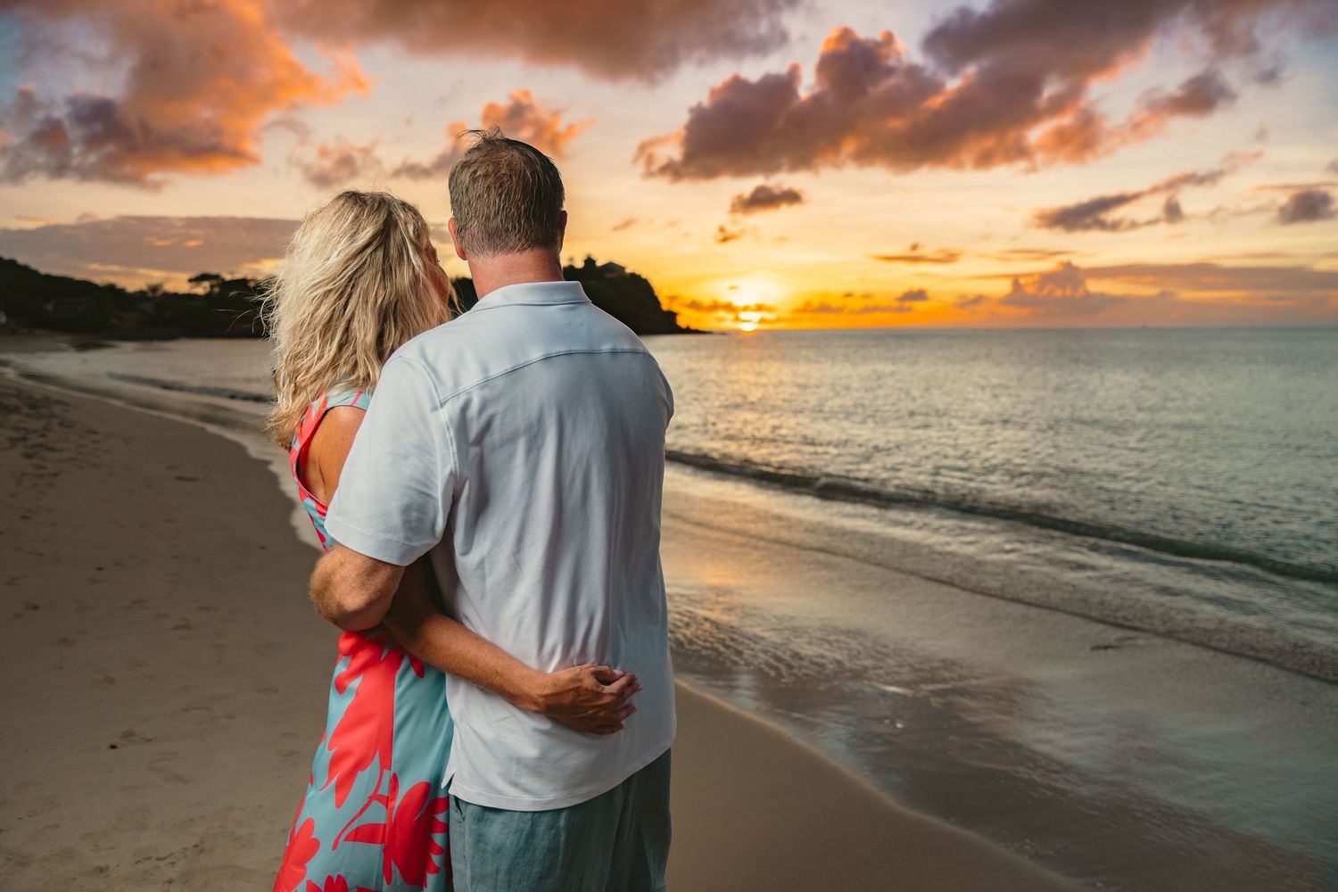 Couple embracing while watching colorful sunset over ocean on tropical sandy beach.