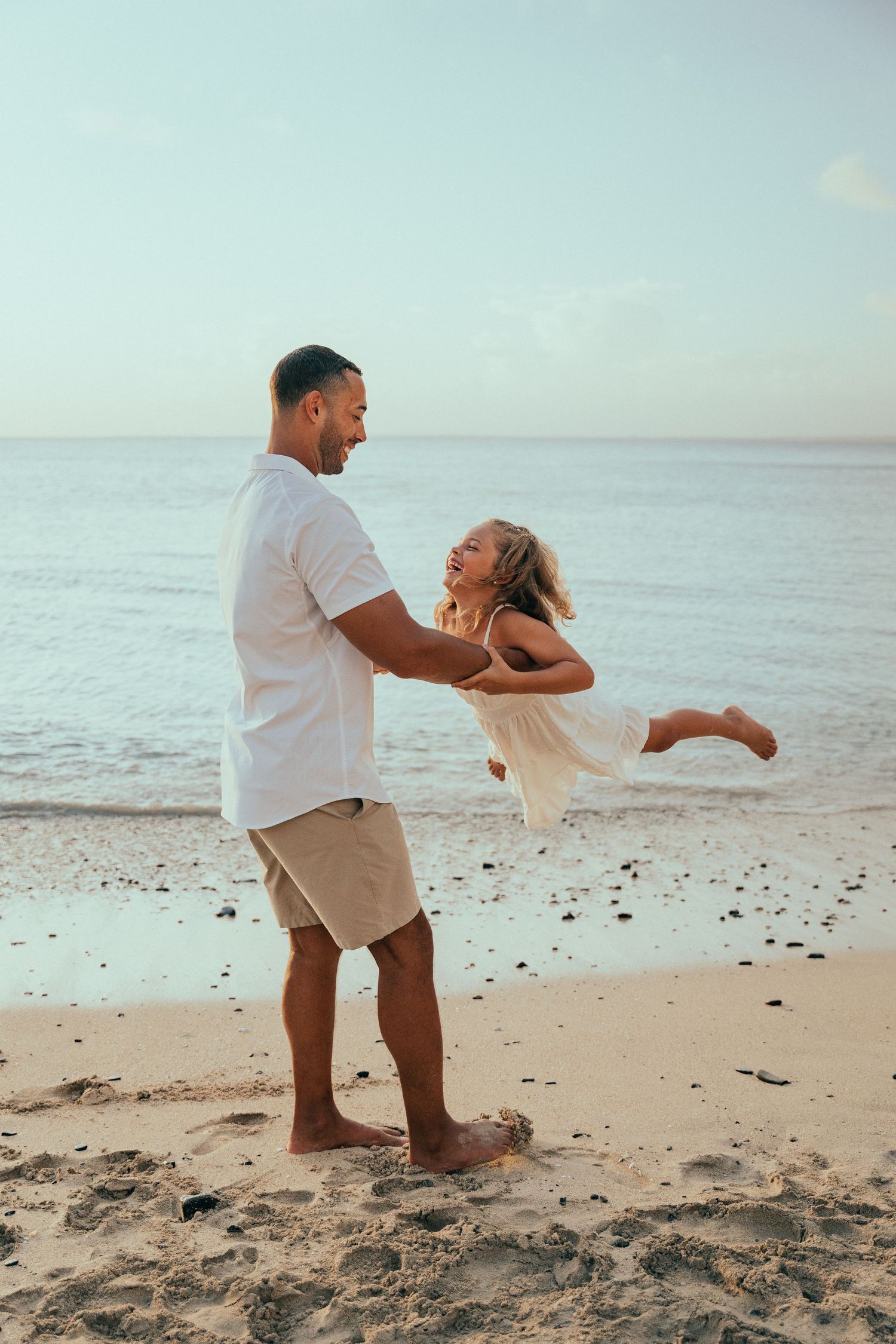 Happy father spinning laughing daughter on a sandy beach with ocean in the background.