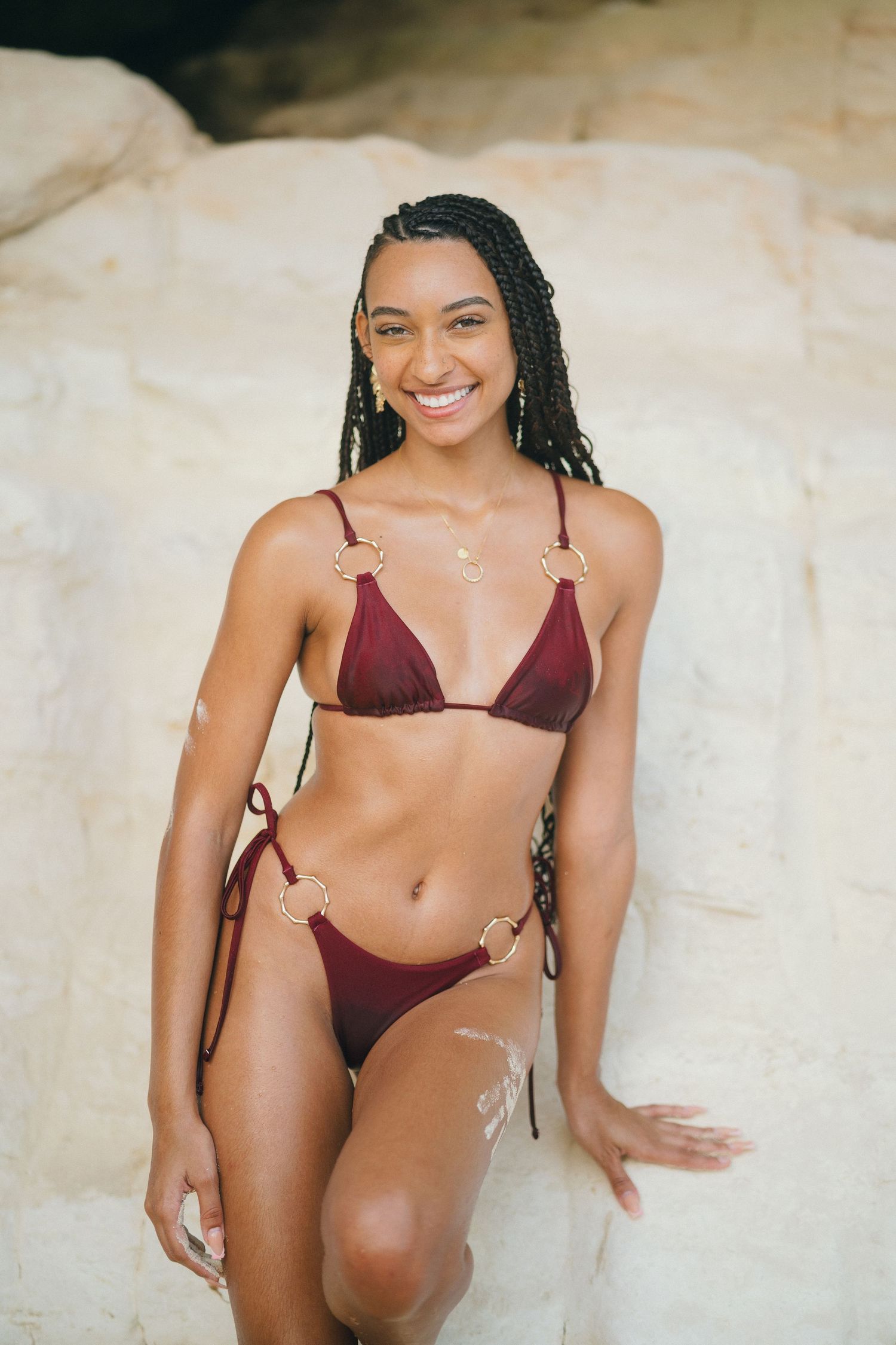 Smiling young woman with braids wearing a burgundy triangle bikini posing against white rock formations.
