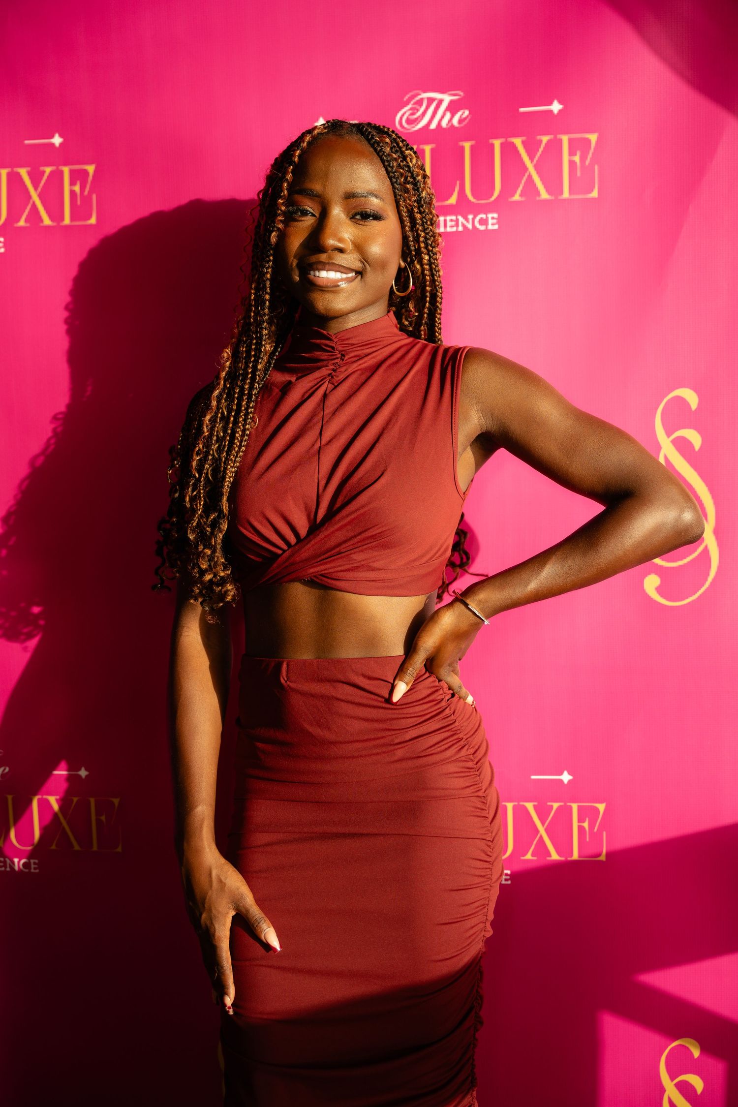 Woman in burgundy two-piece outfit posing at The Luxe Experience event on pink backdrop.