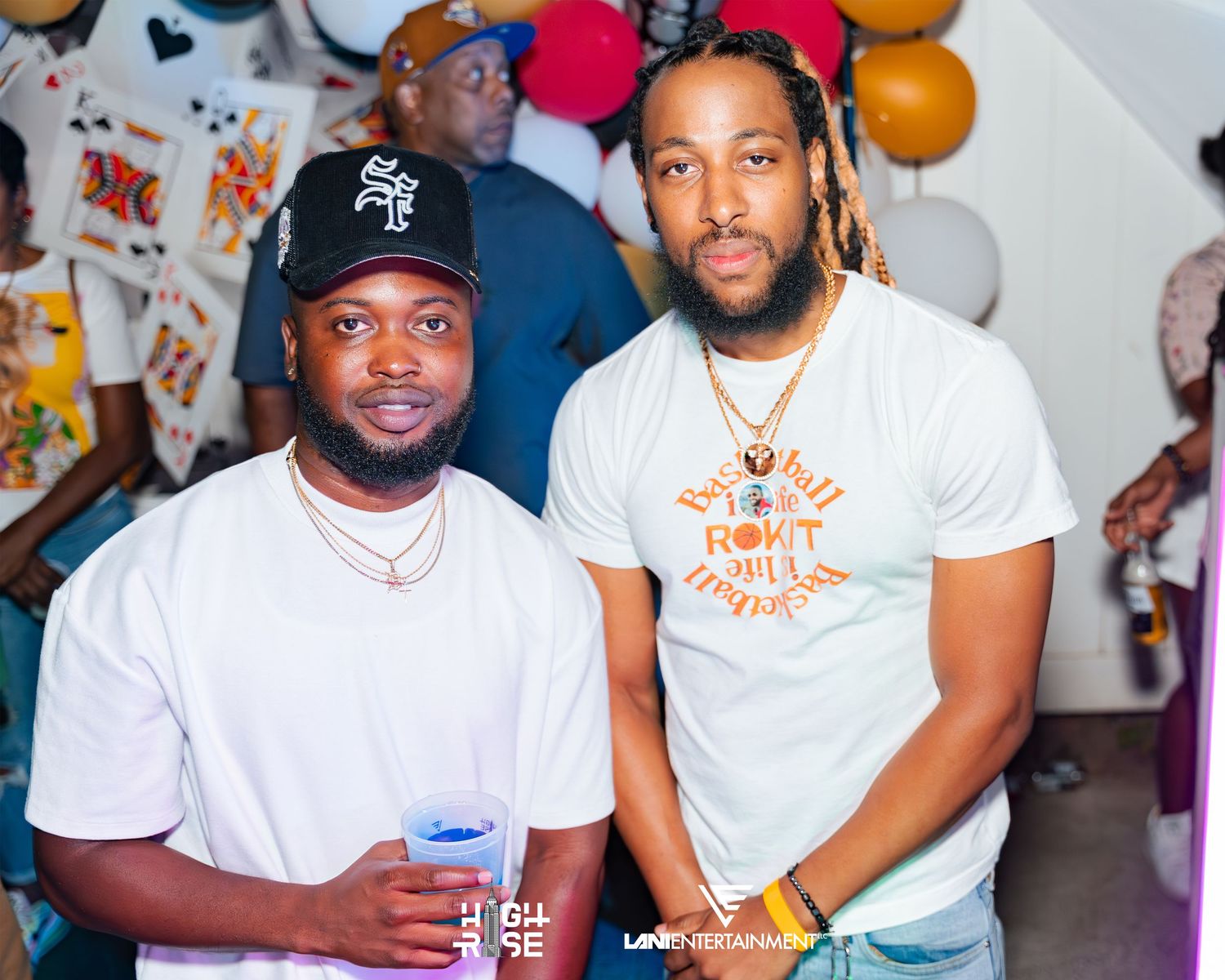 Two men posing together at a colorful party event, both wearing white shirts and casual accessories.