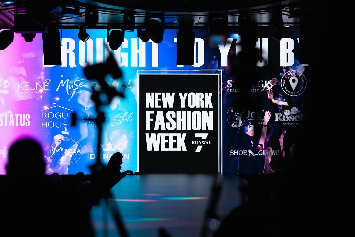 New York Fashion Week runway show with colorful neon signs and silhouettes of photographers in the foreground.