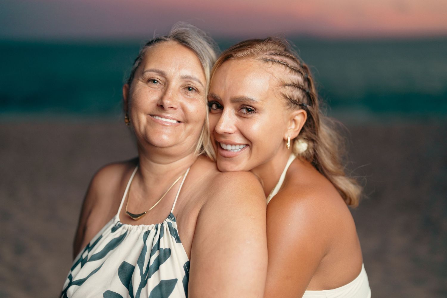 Two smiling women pose together on a beach at sunset.