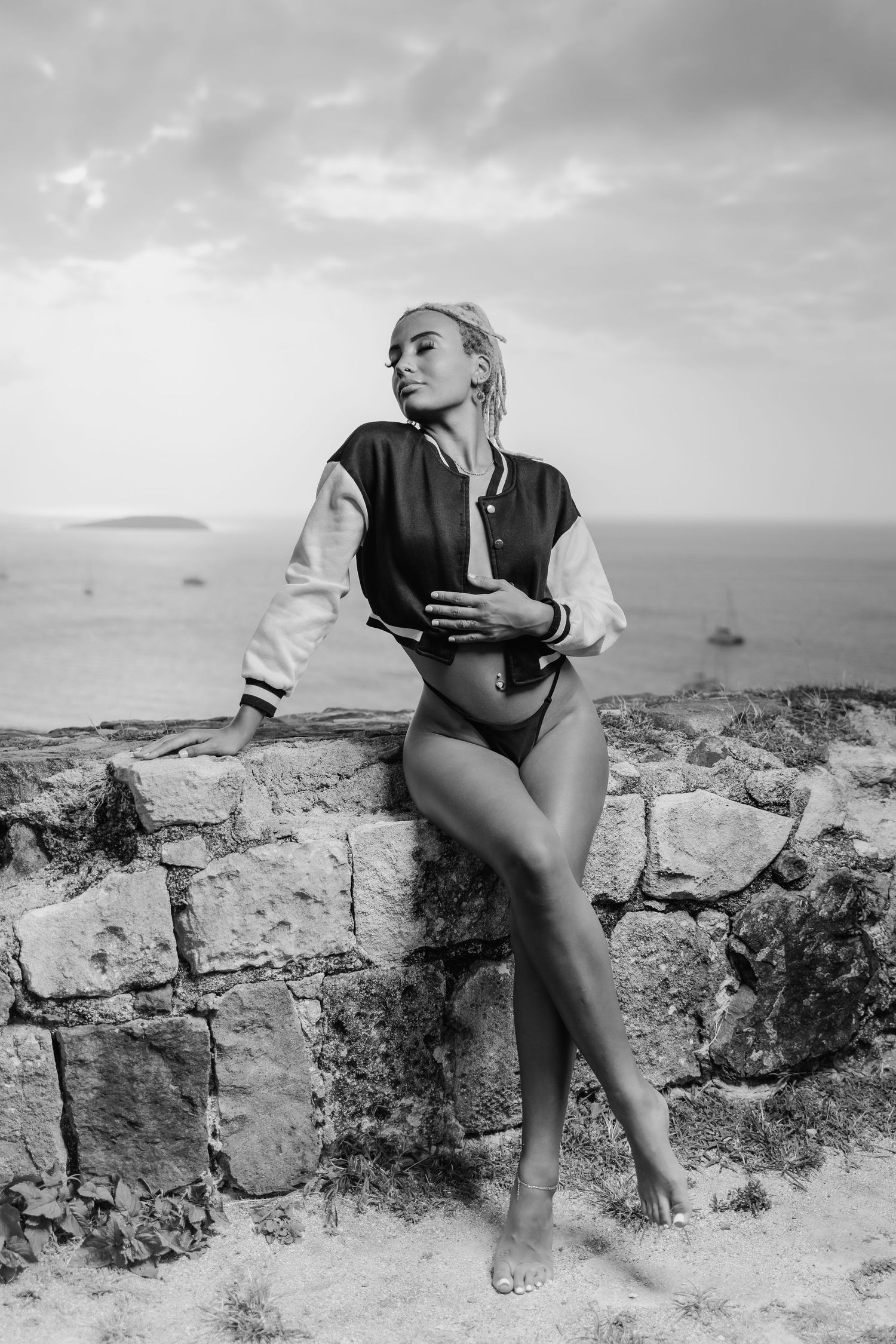 Black and white photo of a blonde woman in a varsity jacket and bikini posing on stone wall by the sea.