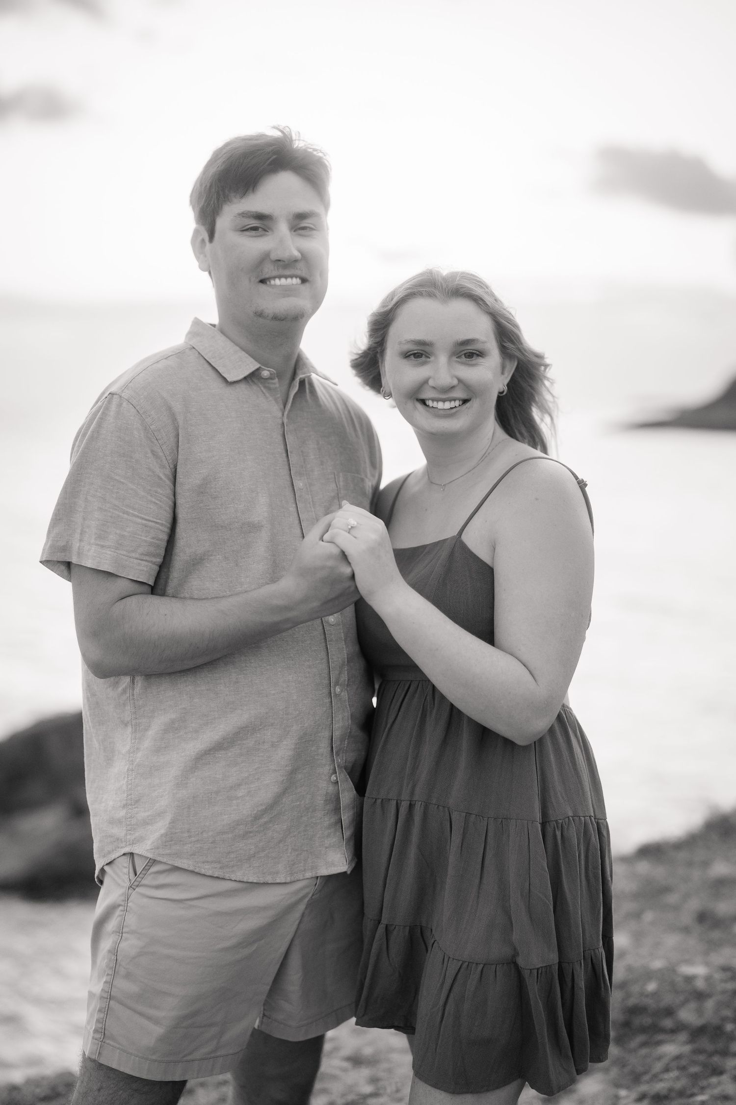 Smiling couple holding hands on a beach at sunset in black and white engagement photo.