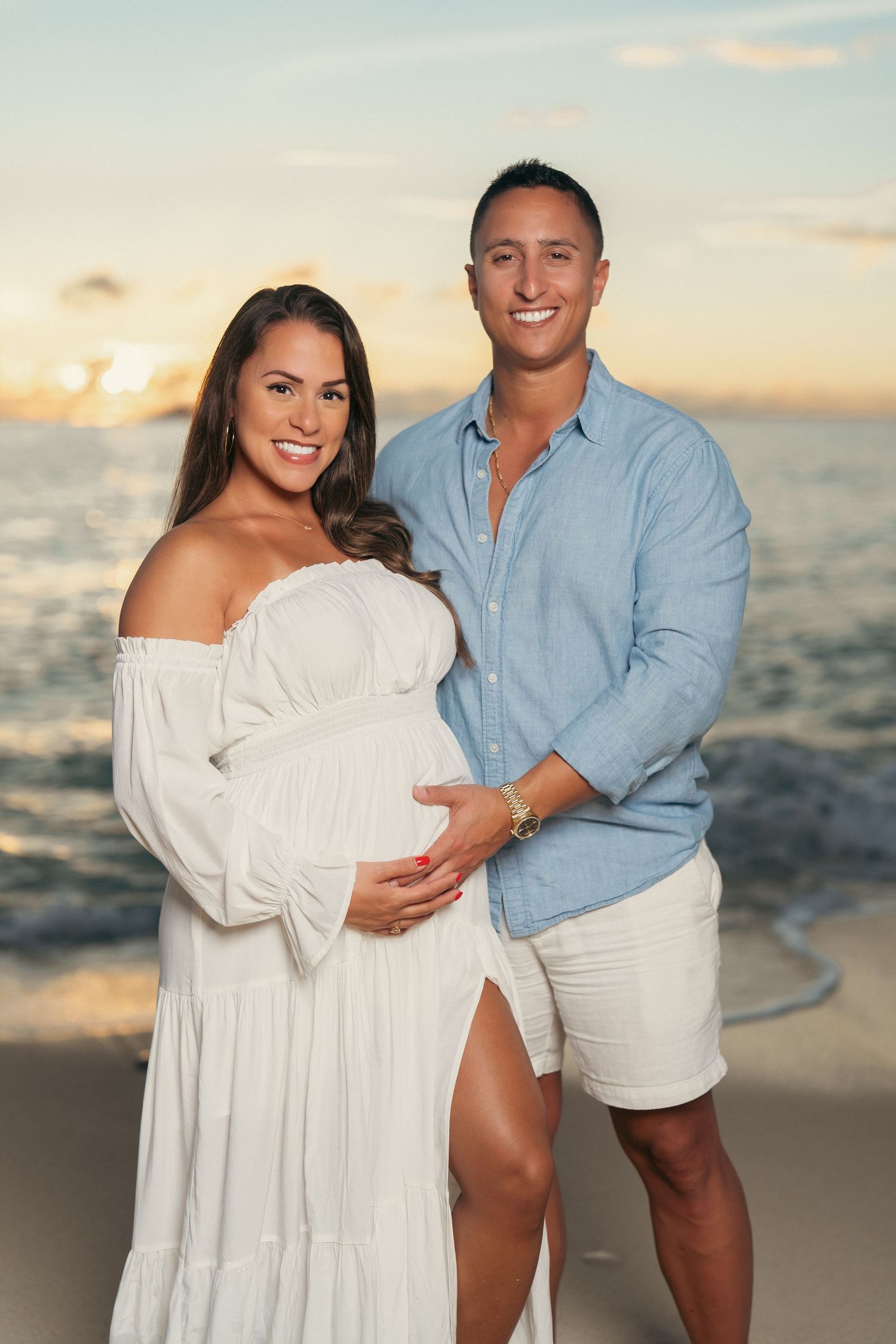 Smiling couple on beach at sunset, pregnant woman in white dress with partner in blue shirt.
