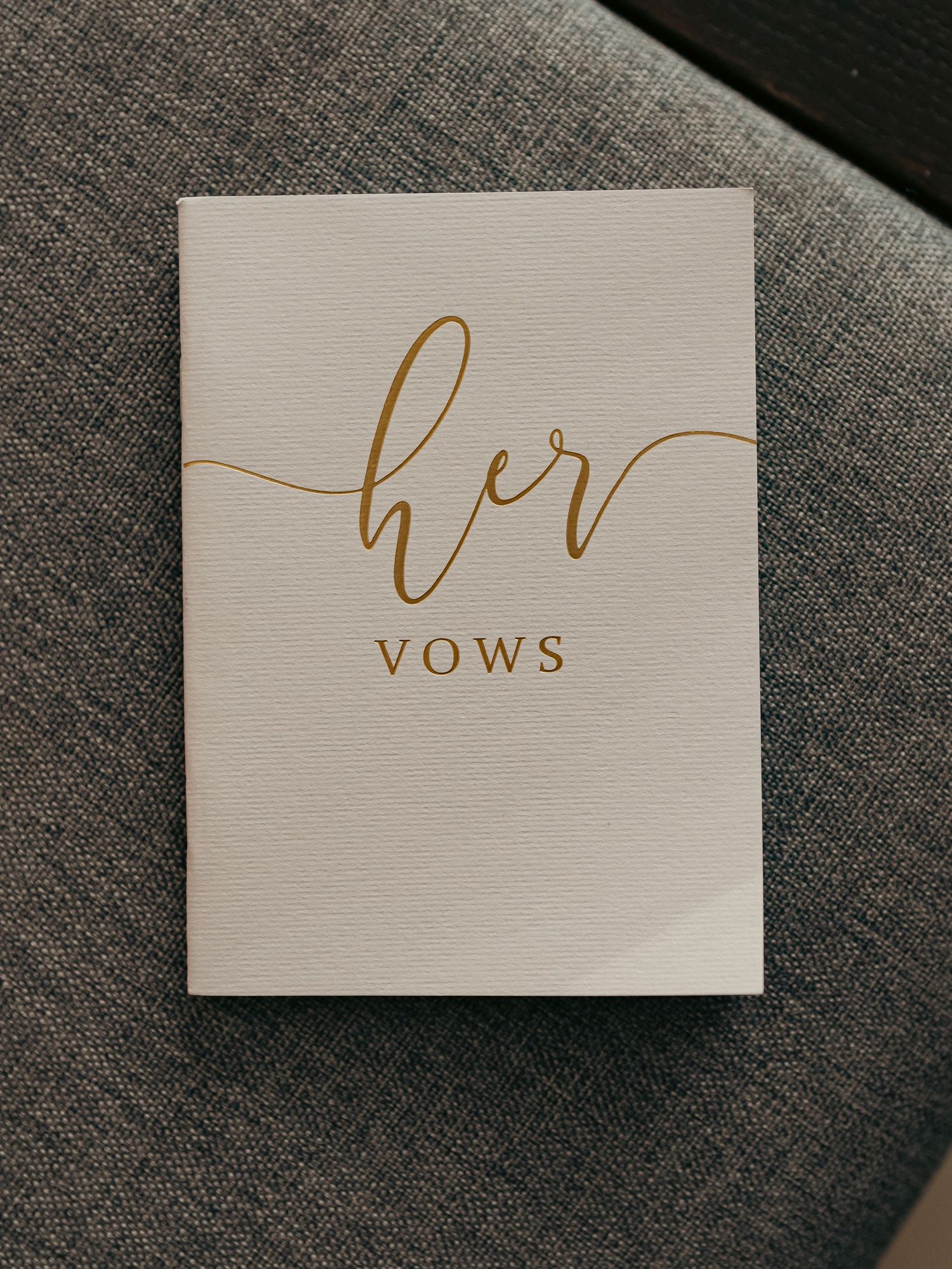 Elegant white wedding vow booklet with gold script reading her vows resting on a gray fabric surface.