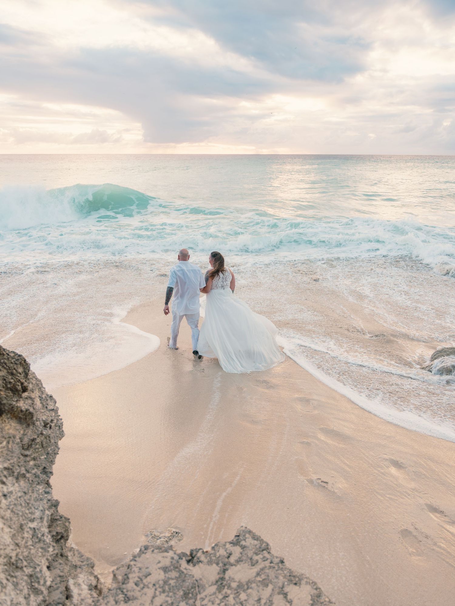 Bride in flowing white gown and groom in white suit walking hand-in-hand along a dramatic wave-swept beach at sunset.