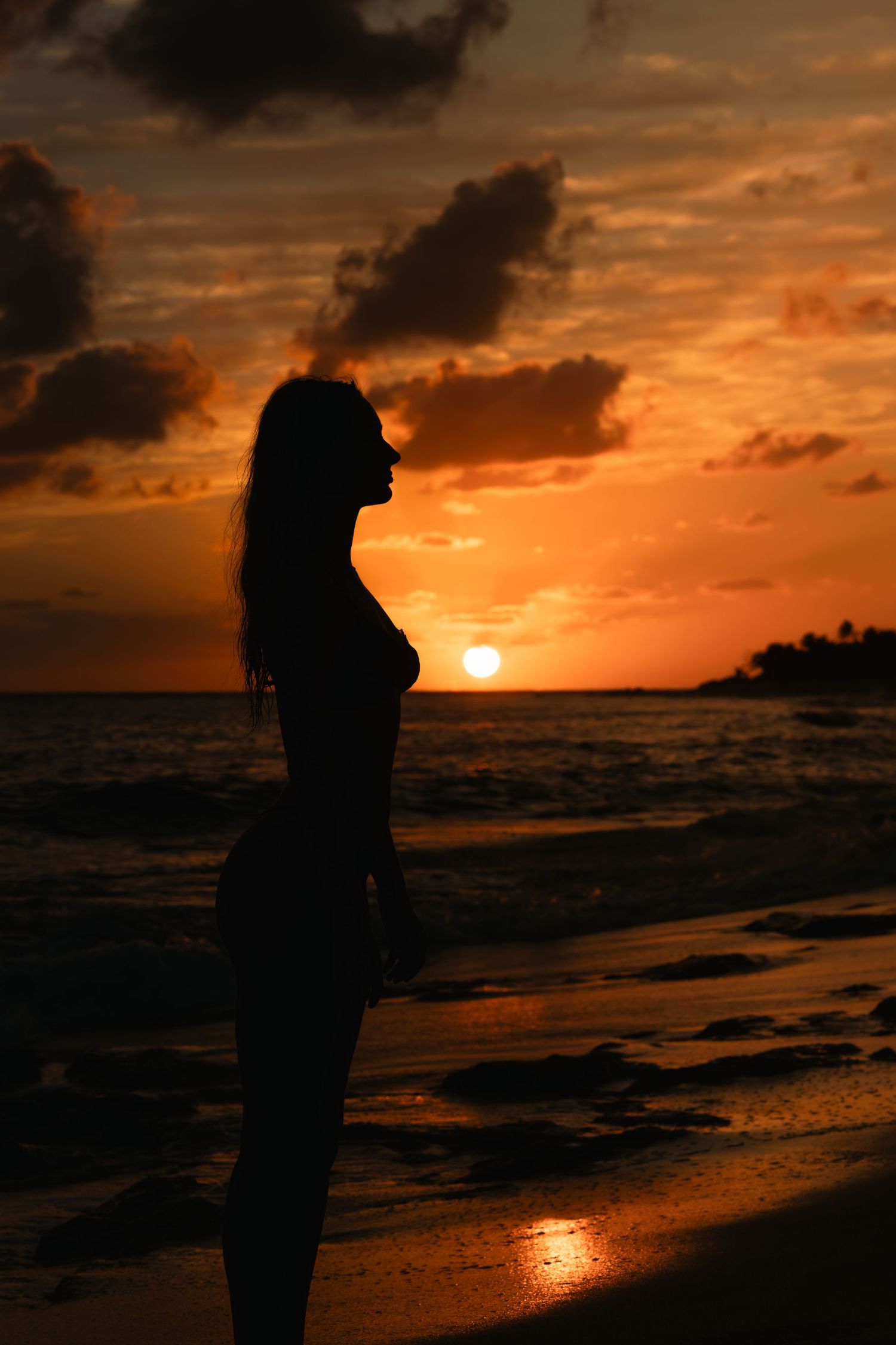 Silhouette of woman standing on beach shore during dramatic orange sunset.