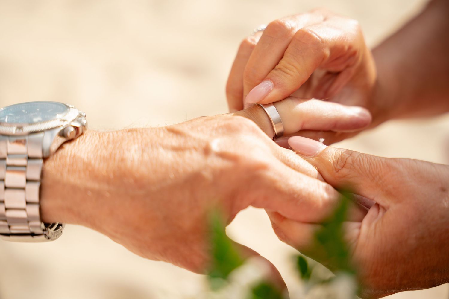 Close-up of couple exchanging wedding rings on sandy beach with groom wearing silver watch.