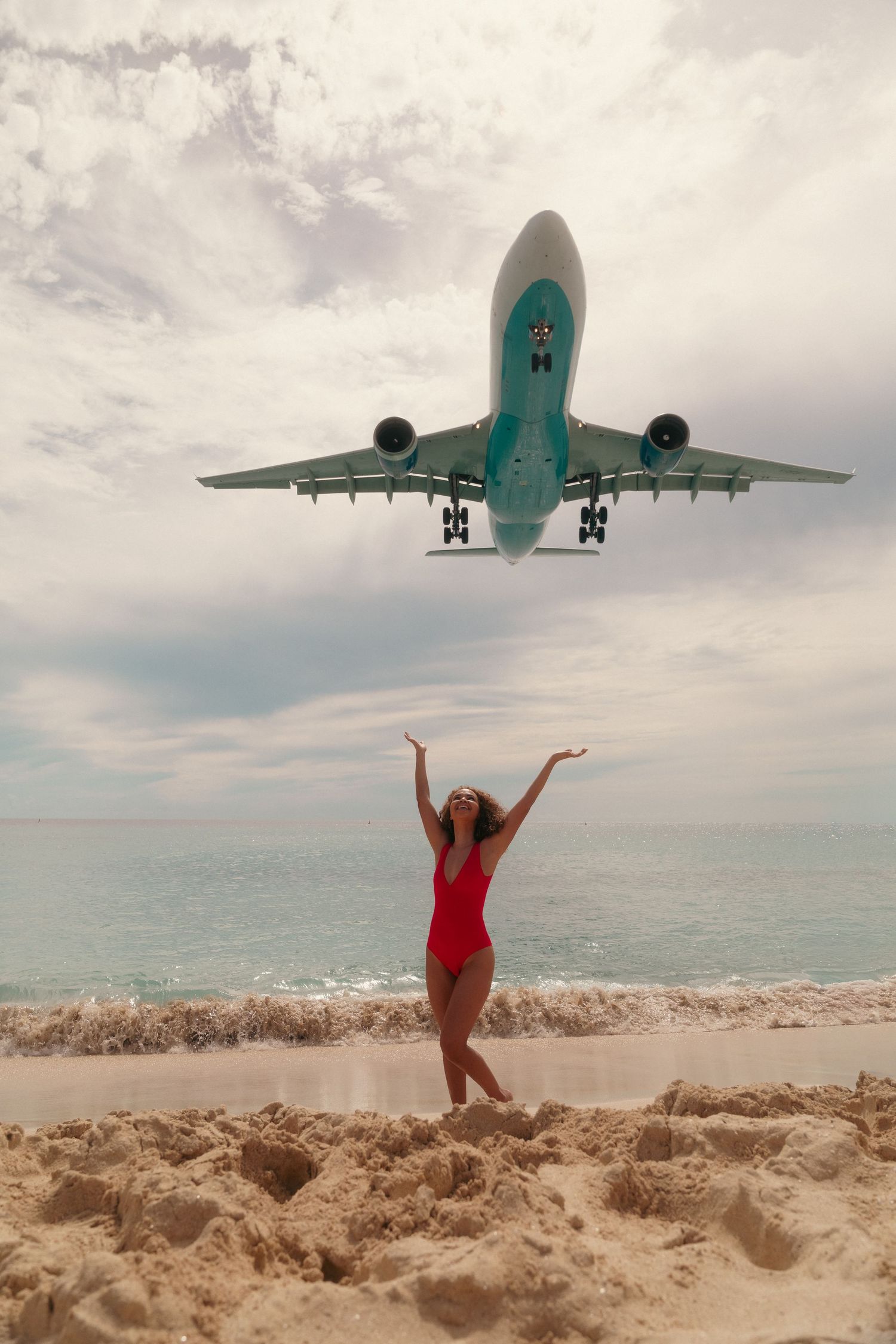Woman in red swimsuit on beach with arms raised as large airplane flies overhead.