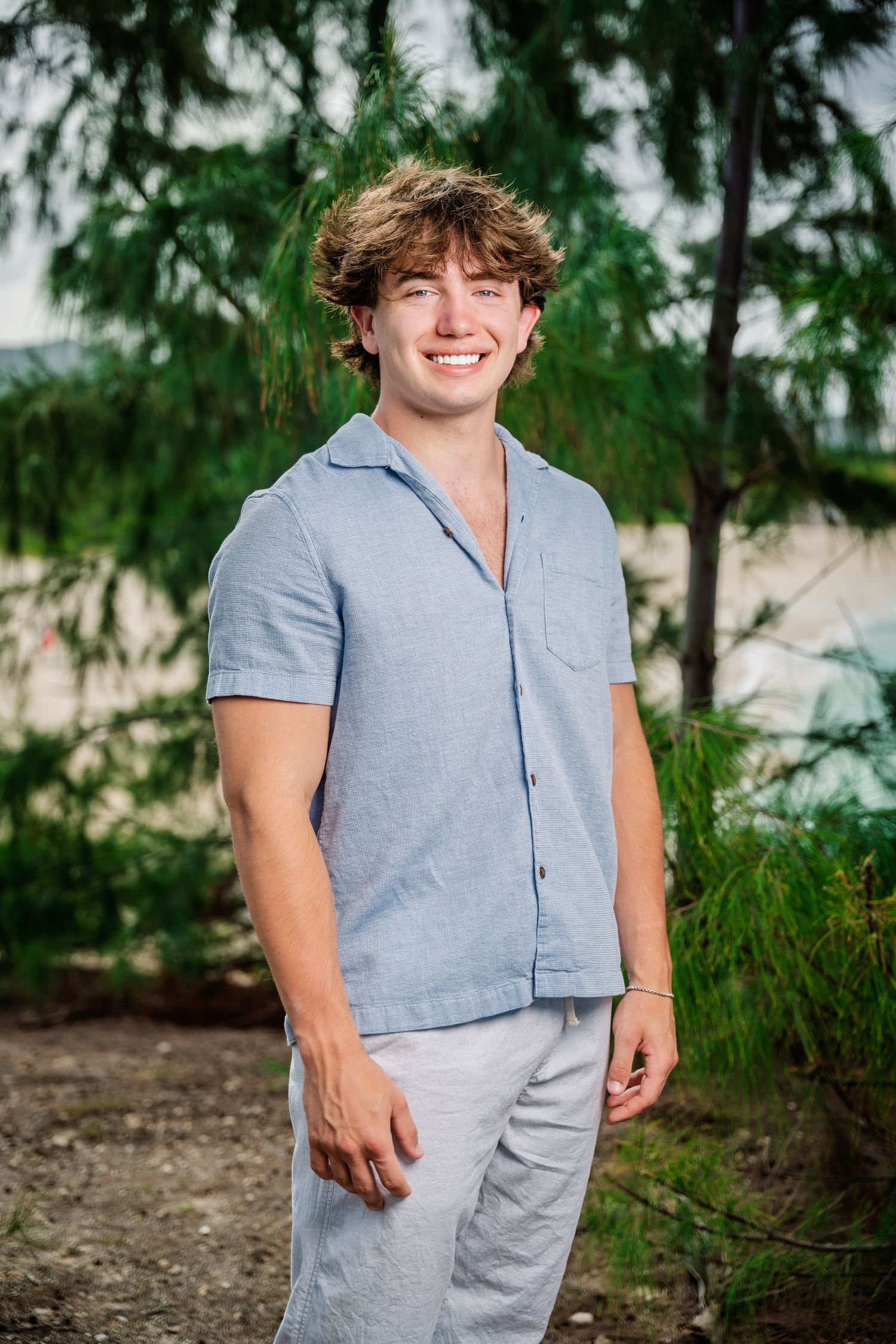Happy young man with curly hair in a blue button-up shirt smiling outdoors near tropical pine trees.