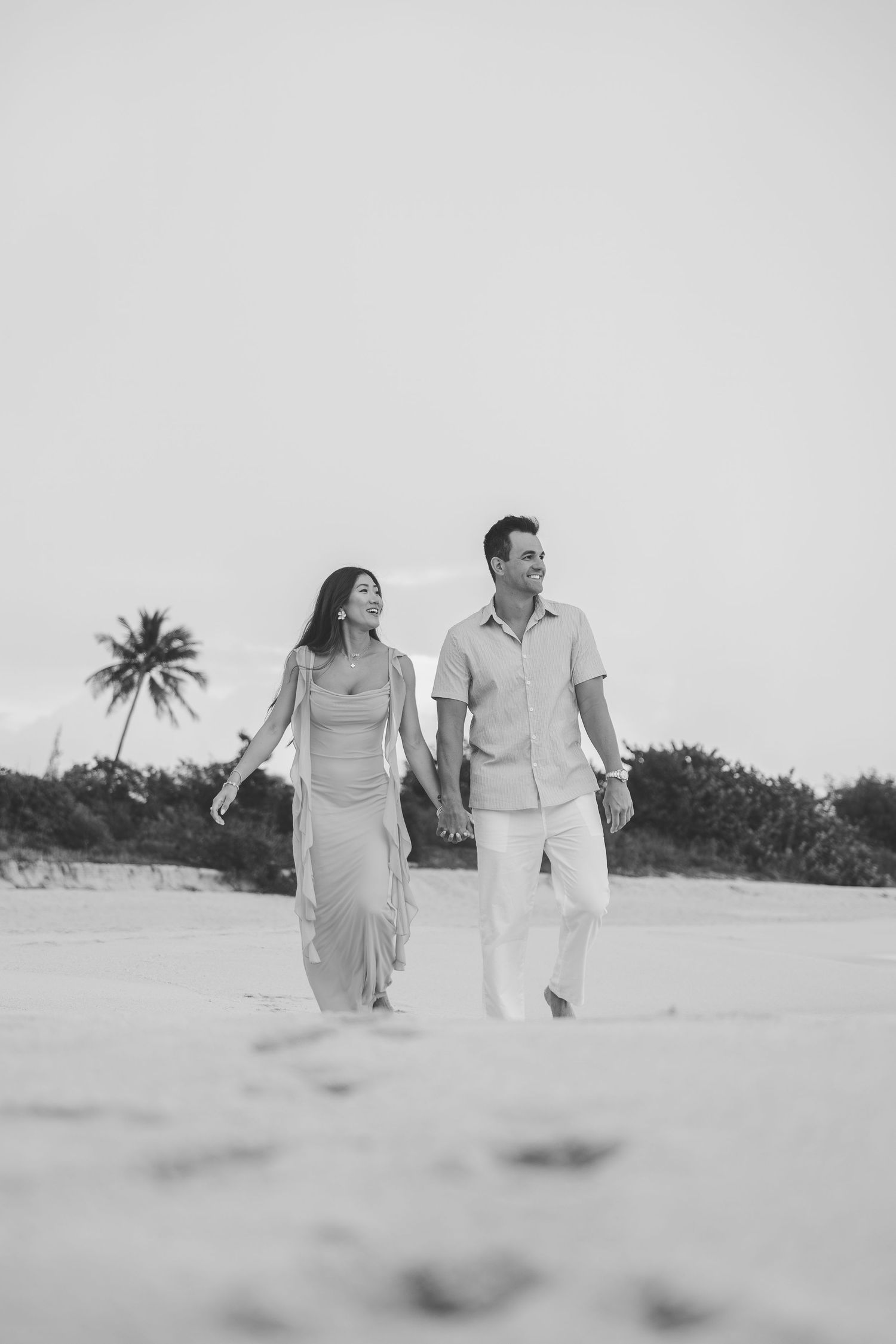 Black and white photo of couple holding hands walking on sandy beach with palm tree.
