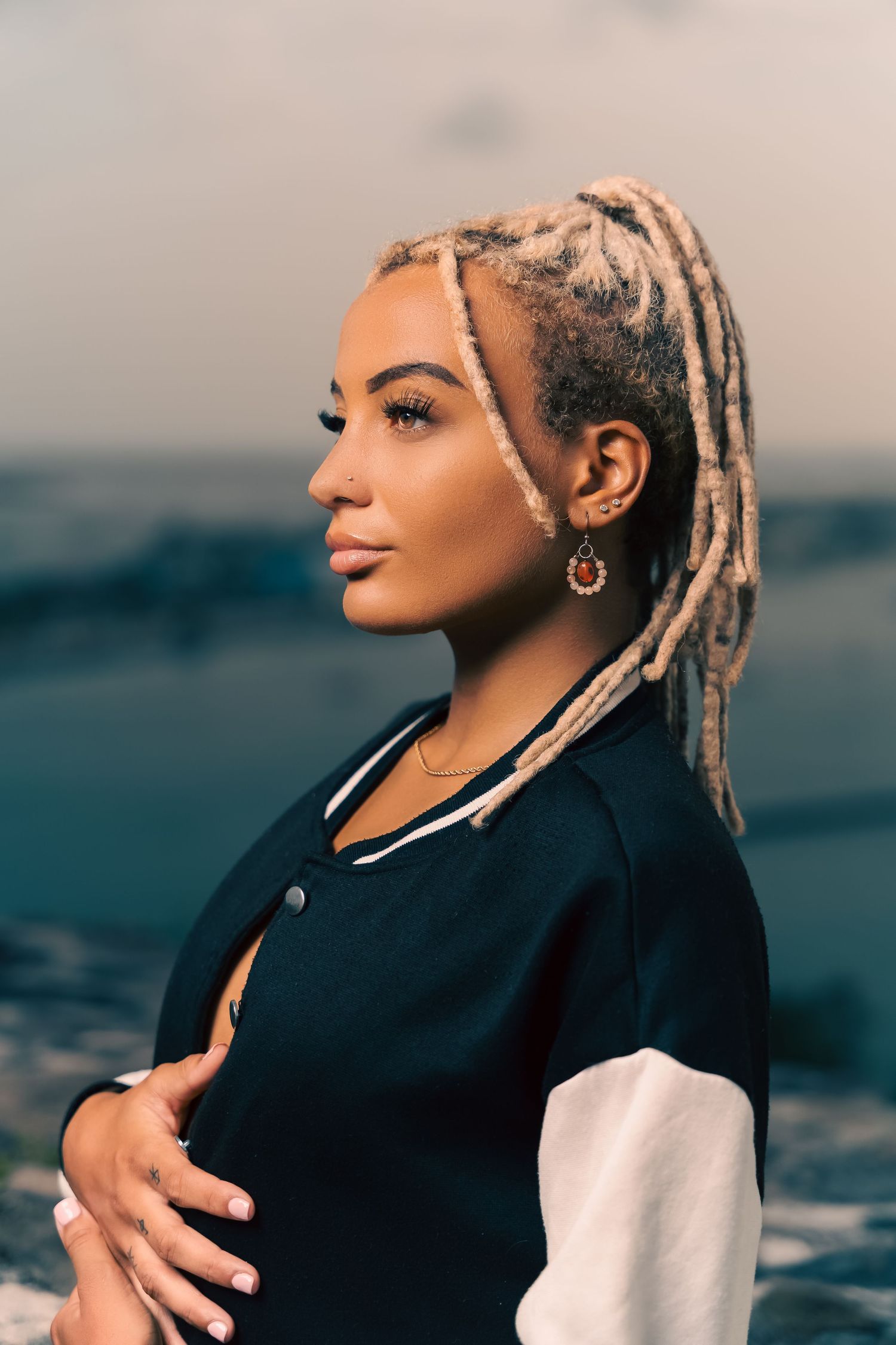 Young woman with blonde dreadlocks wearing a varsity jacket poses outdoors with ocean view at sunset.