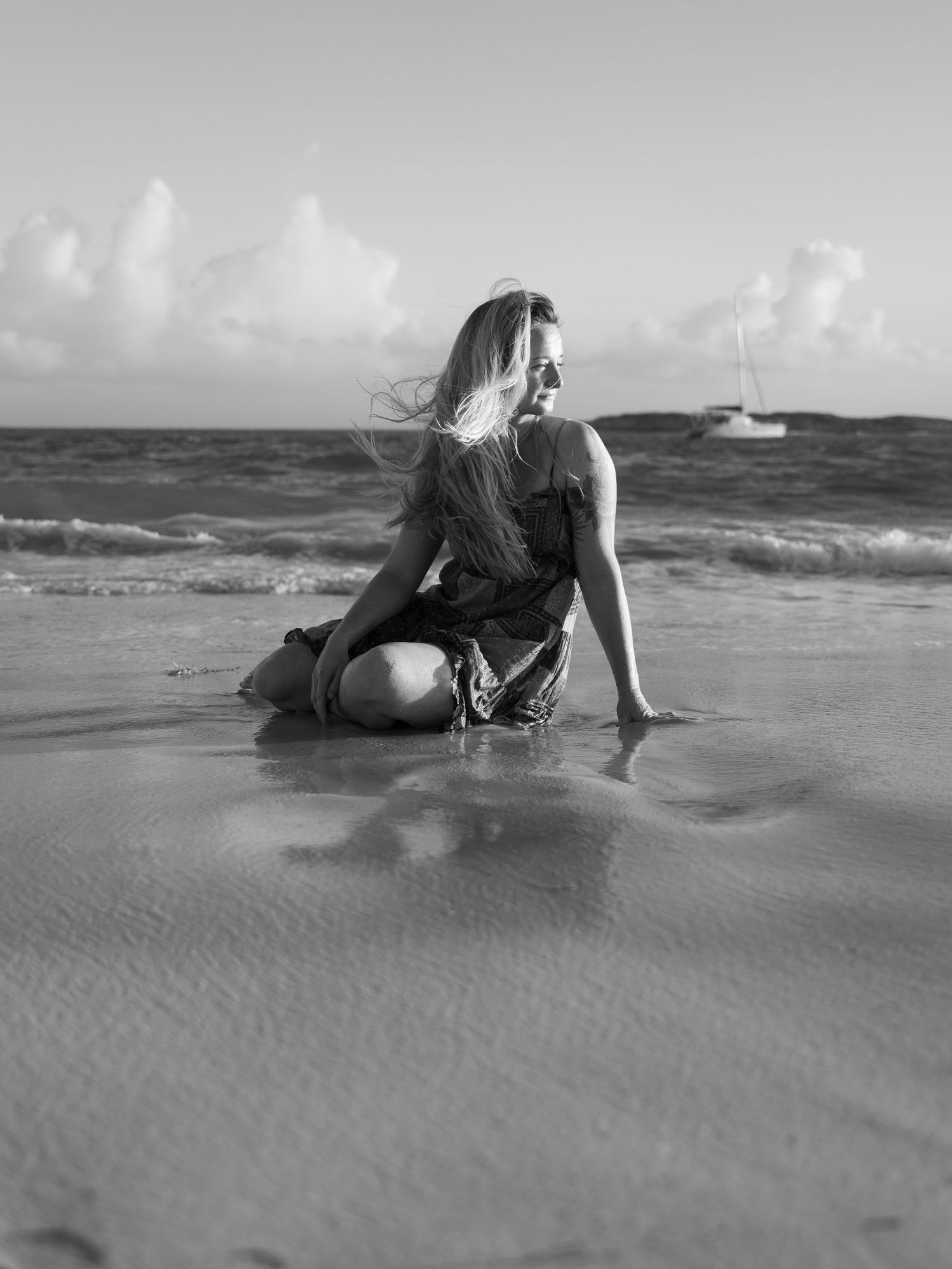 Black and white image of a woman kneeling on wet sand at the beach with a sailboat in the distance.