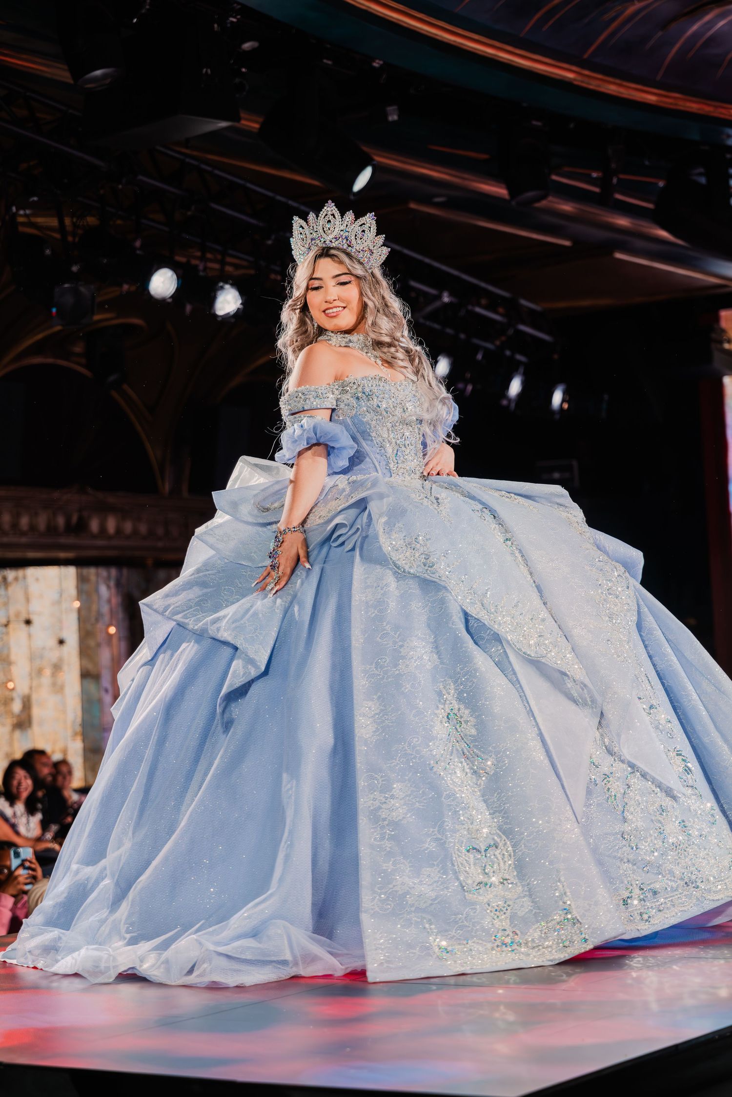 Young woman in a light blue ball gown with silver embroidery and crystal crown walks a runway.