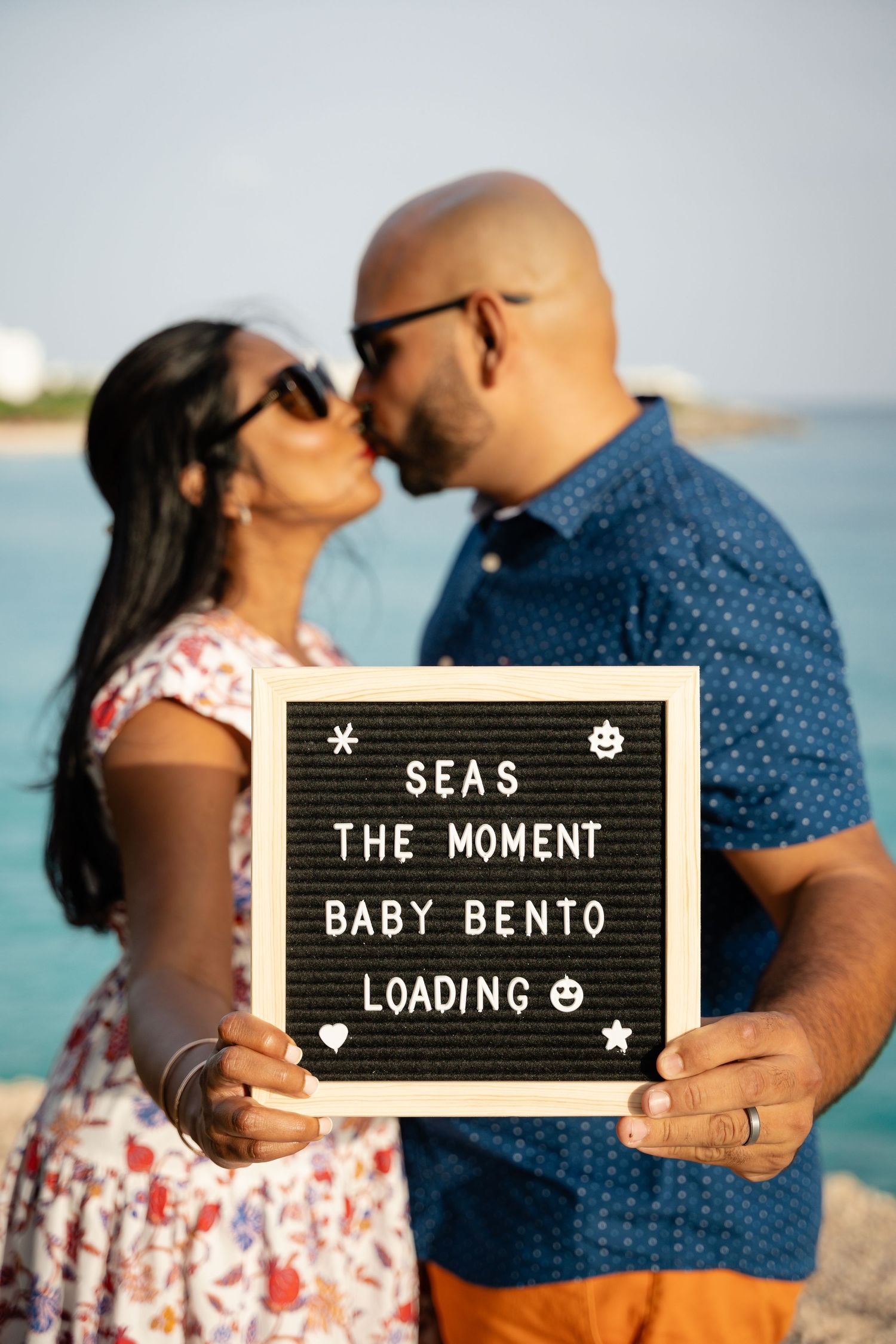 Couple kissing on beach holding letter board reading Seas The Moment Baby Bento Loading.