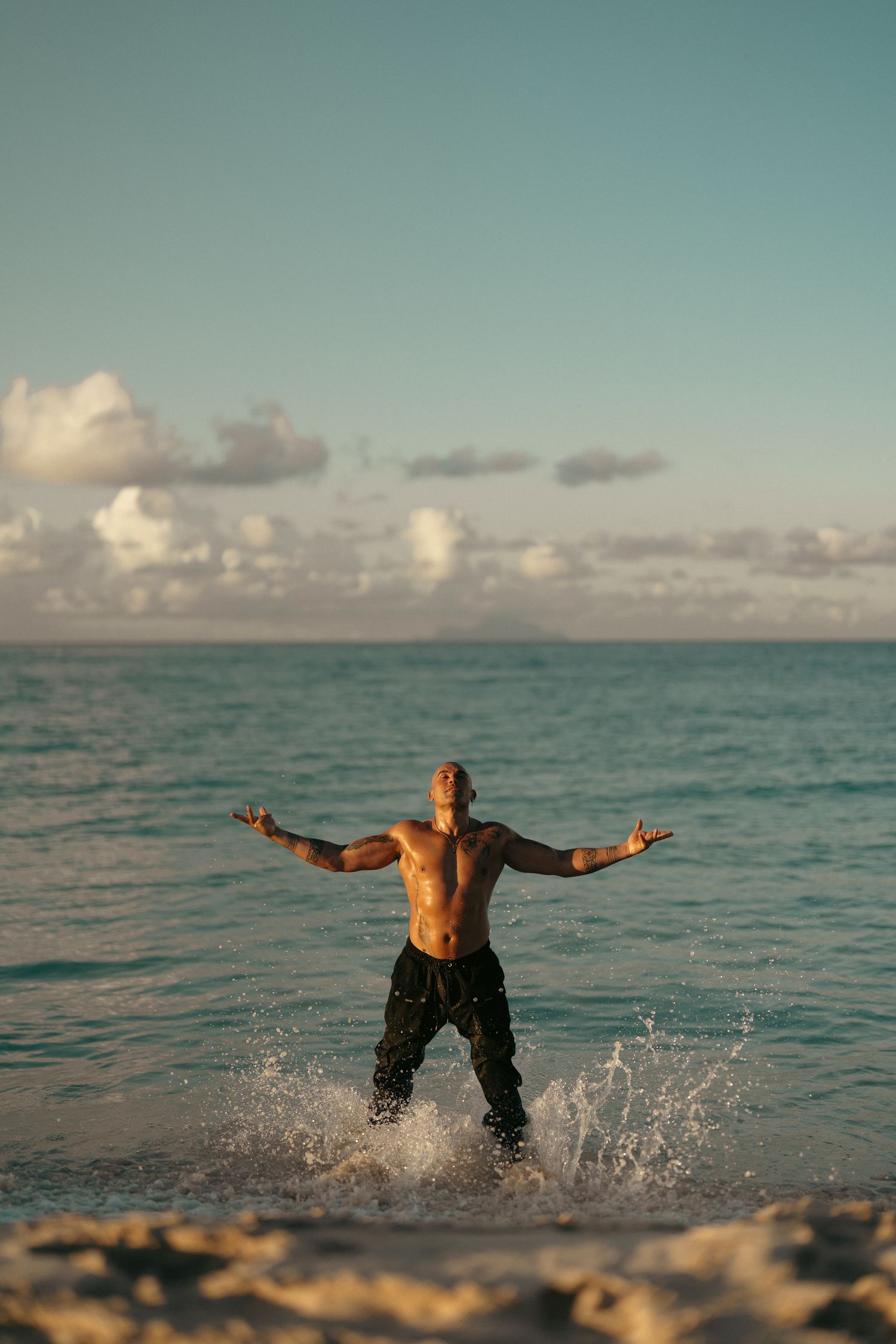 A shirtless muscular man stands with arms outstretched in ocean waves at sunset on a beach.