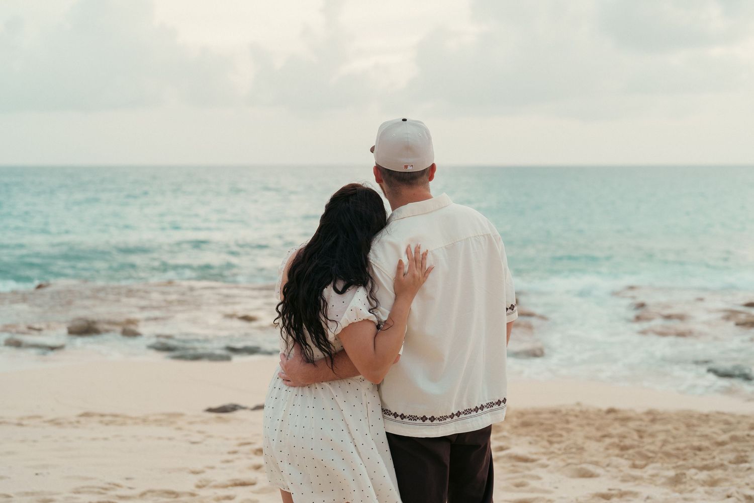 Couple standing arm in arm gazing at ocean waves during beach engagement session.