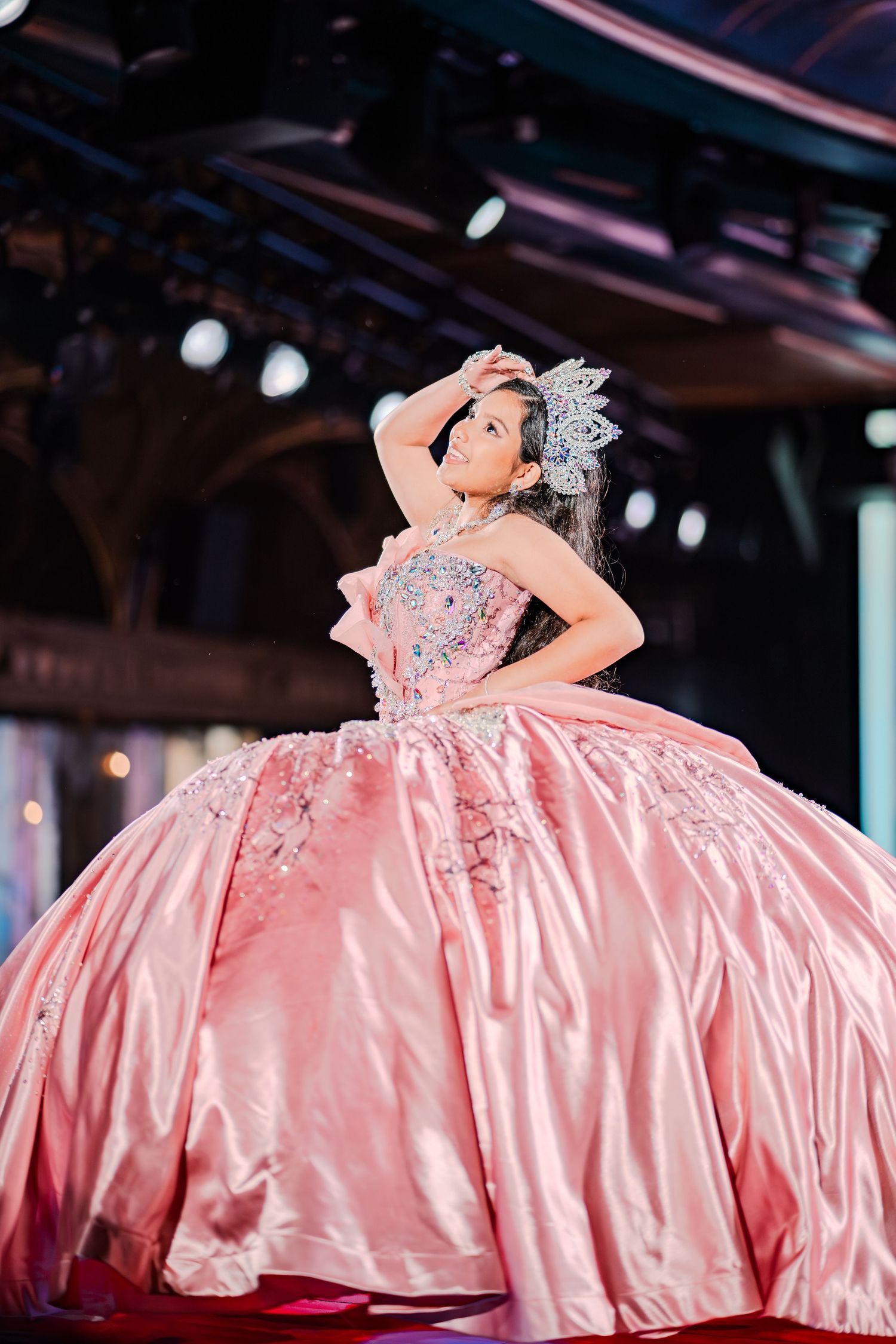 Young girl in a pink ballgown and tiara poses gracefully on stage during a pageant competition.