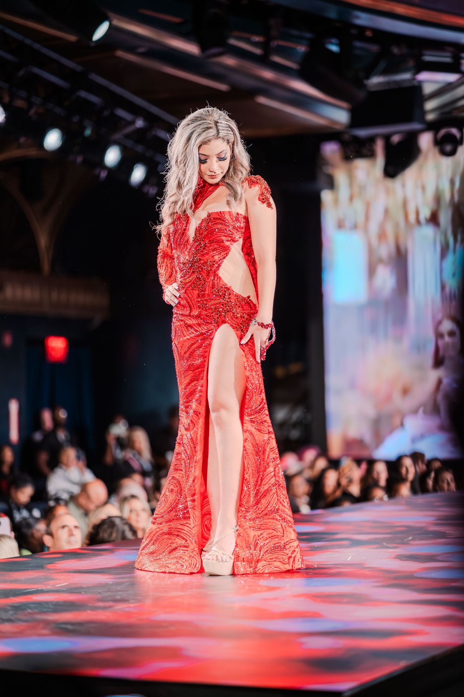 A model walks the runway in a stunning red sequined gown with a high slit and cutout waist at a fashion show.