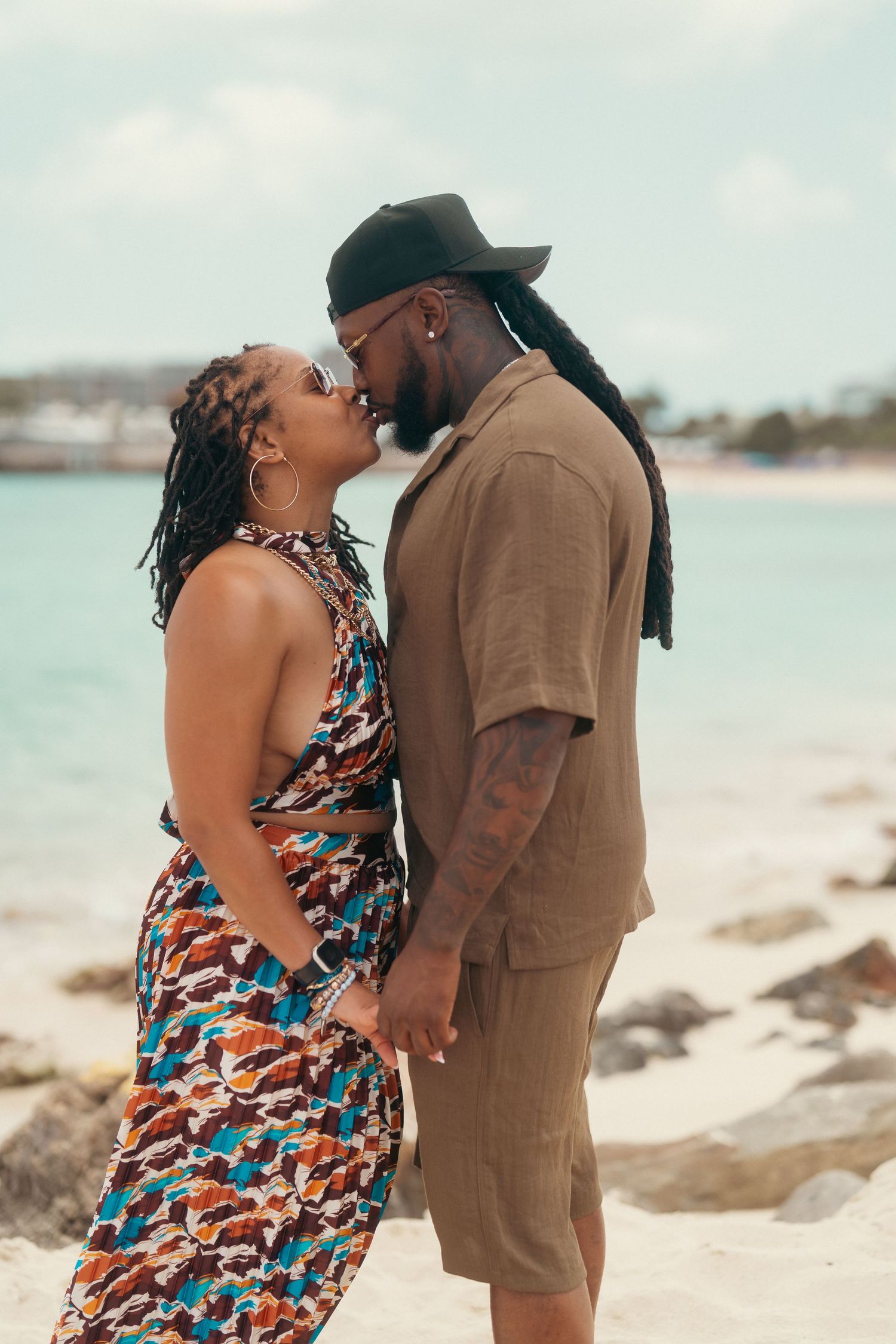 Couple sharing a kiss on beach with colorful outfits and ocean in background.