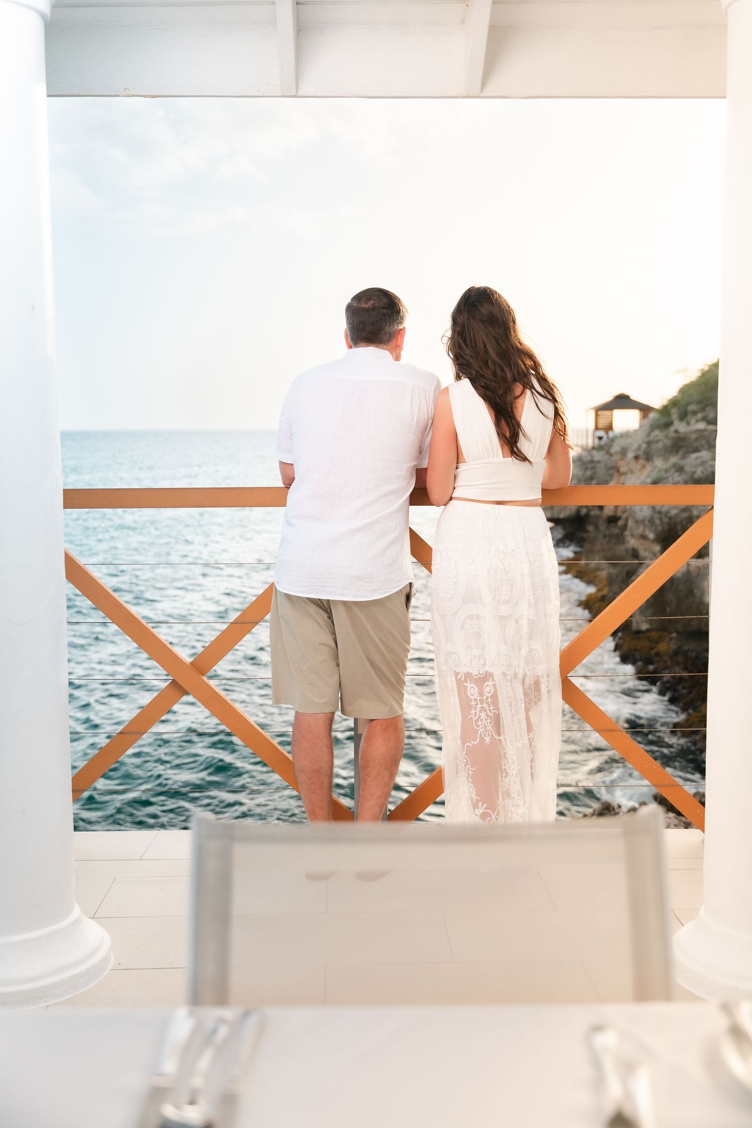 Couple standing together on an ocean-view balcony, woman in lace dress and man in white shirt enjoying the sea view.