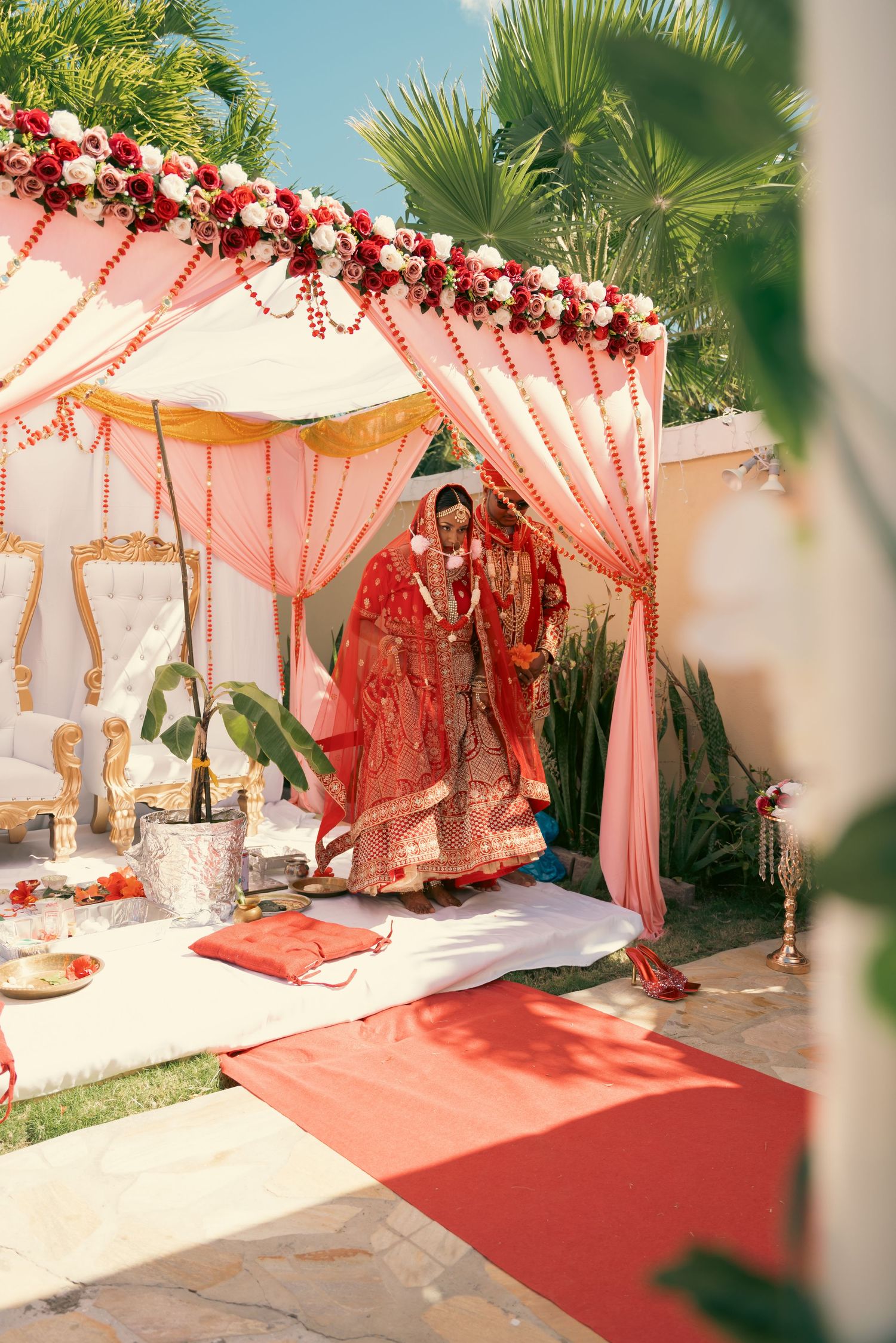 Indian bride in red lehenga standing under floral mandap during outdoor wedding ceremony.