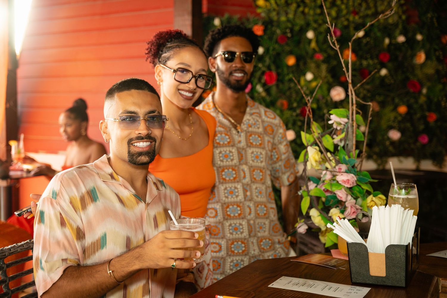 Three friends smiling at an outdoor bar with tropical floral decor, one holding a cocktail.