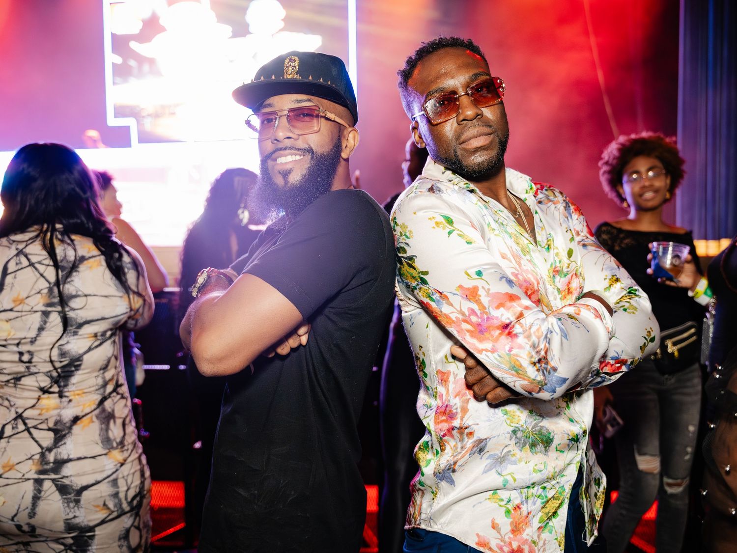 Two men posing with arms crossed at a vibrant indoor music event.