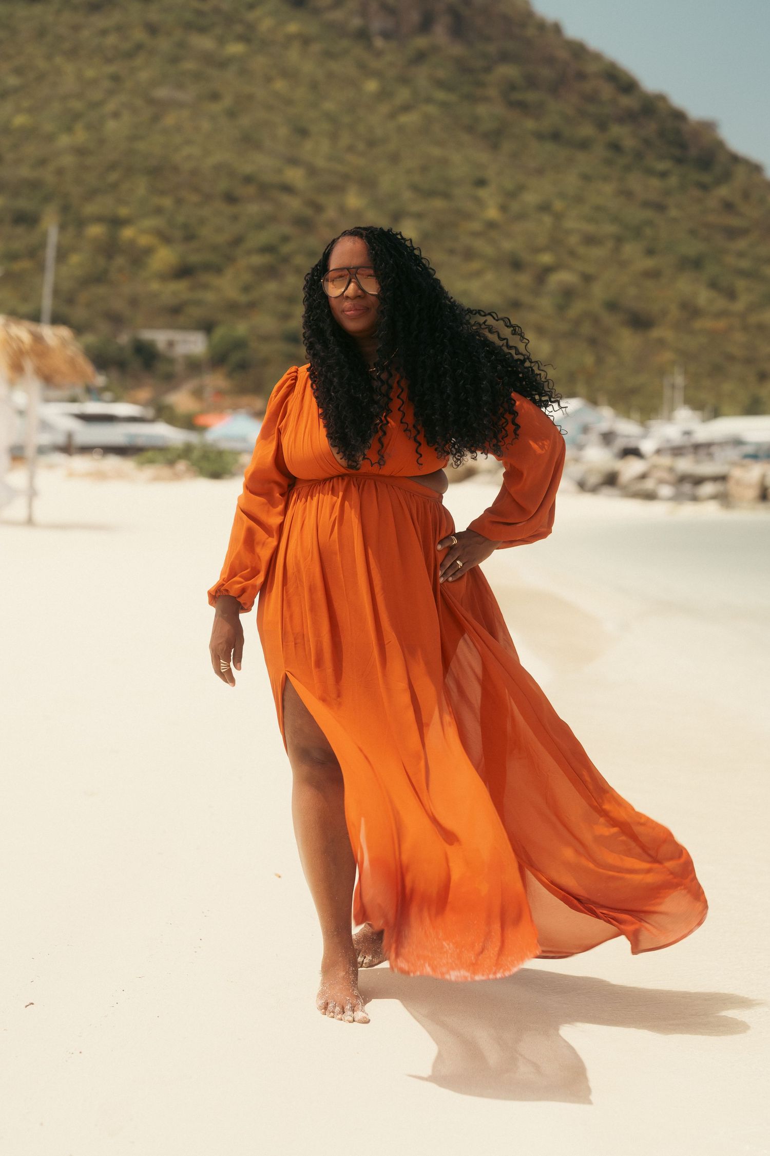 Woman in flowing orange maxi dress walks gracefully on white sand beach with mountain and boats in background.