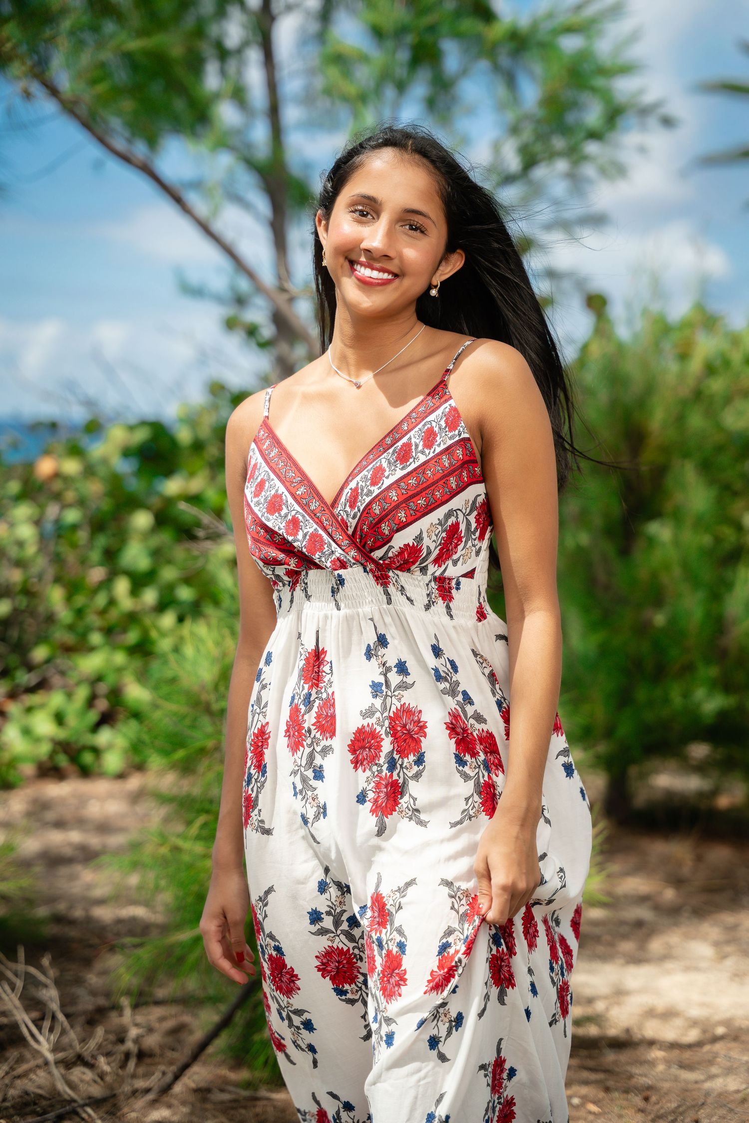 Smiling young woman in floral red and white sundress standing outdoors among tropical green foliage.