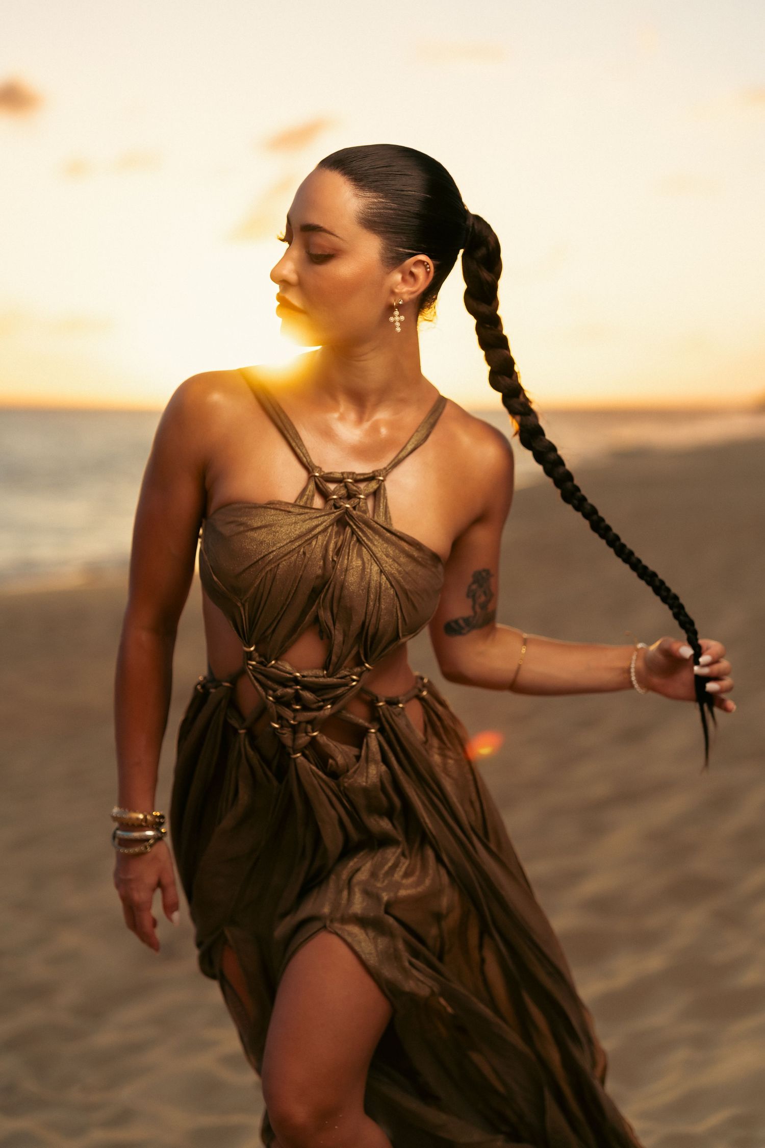 Woman in a gold draped dress holding a long braid while posing on a beach during a golden sunset.