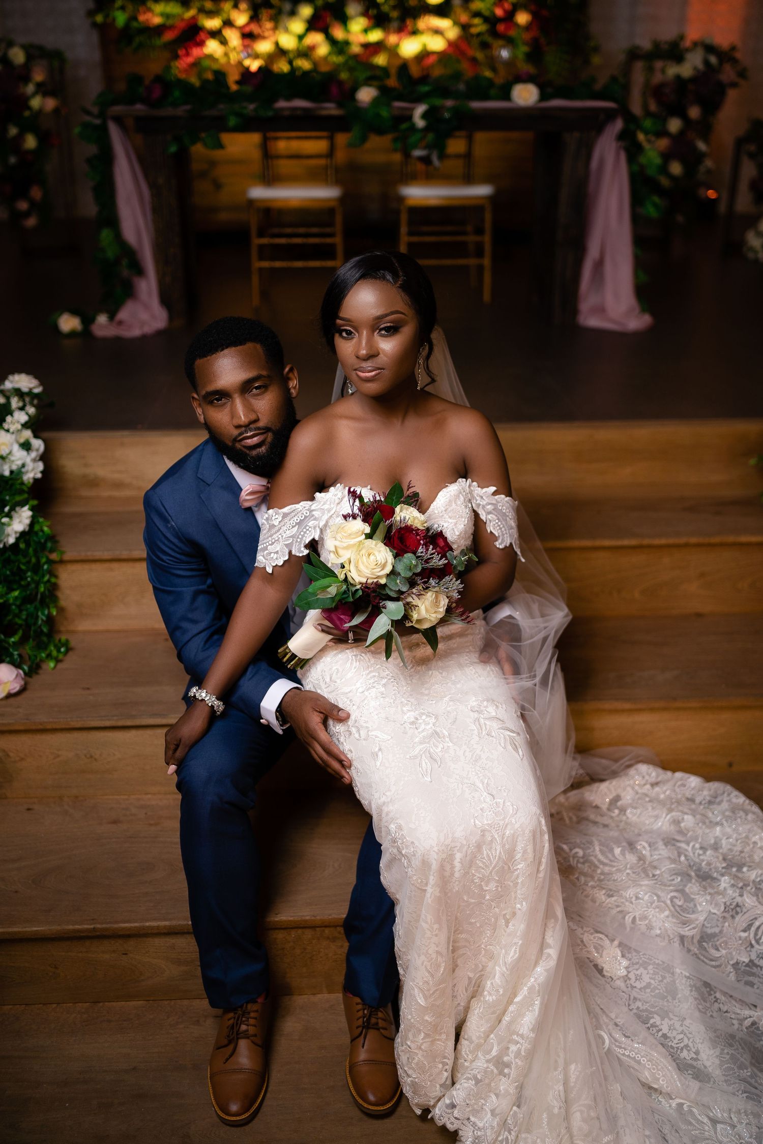 Bride and groom pose together at decorated wedding venue, bride holding roses bouquet.