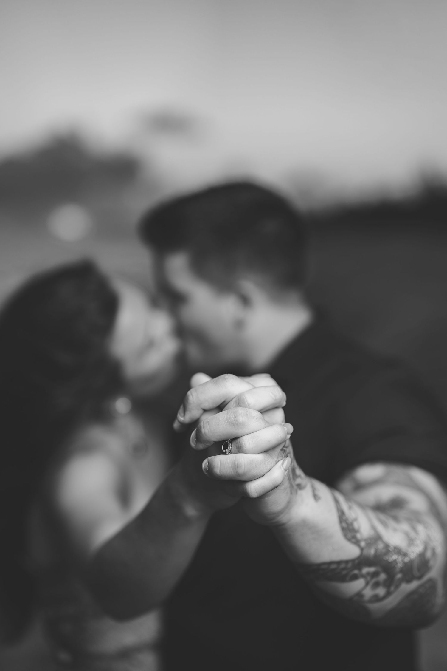 Black and white romantic photo of couple kissing while holding hands, man's arm features a tattoo sleeve.