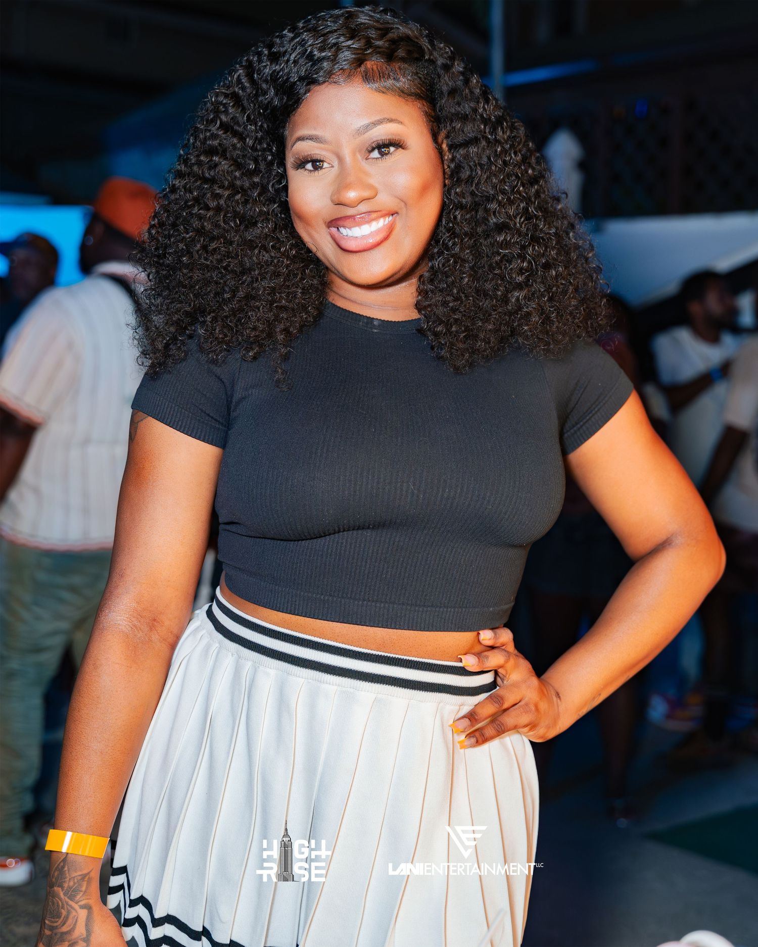 A smiling young woman with curly hair poses in a black crop top and white skirt at a party.
