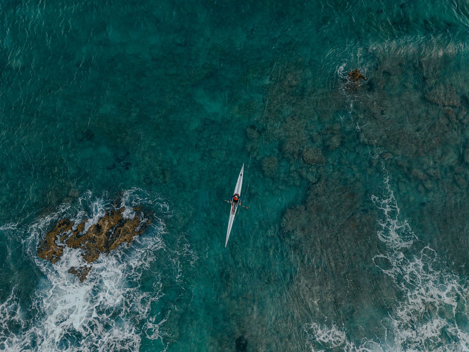Aerial drone view of a lone kayaker paddling through turquoise ocean waters near rocky reef.