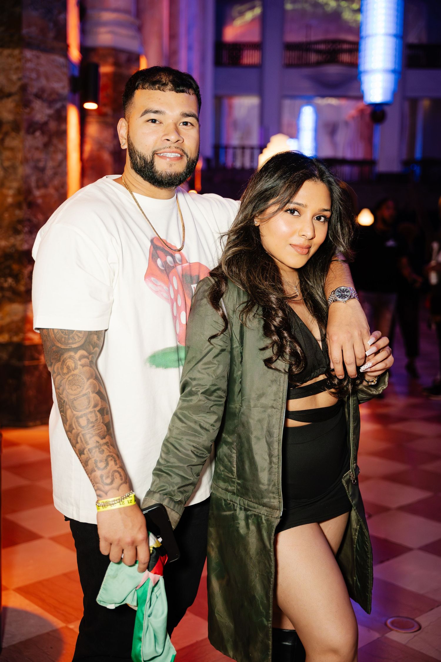 Tattooed man and woman pose together at a colorfully lit indoor event.