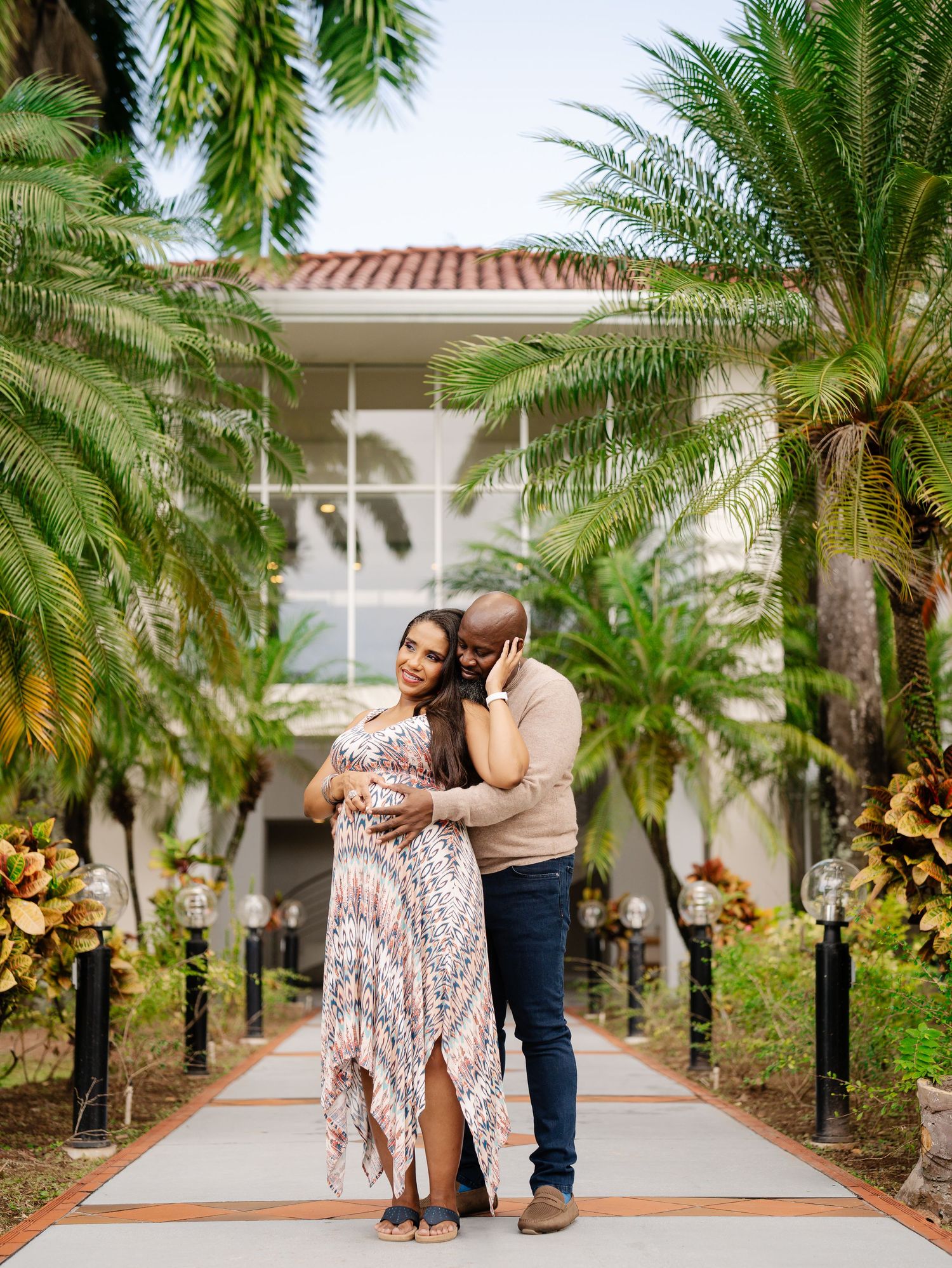 Couple embraces on tropical palm-lined walkway during maternity photo session outdoors.